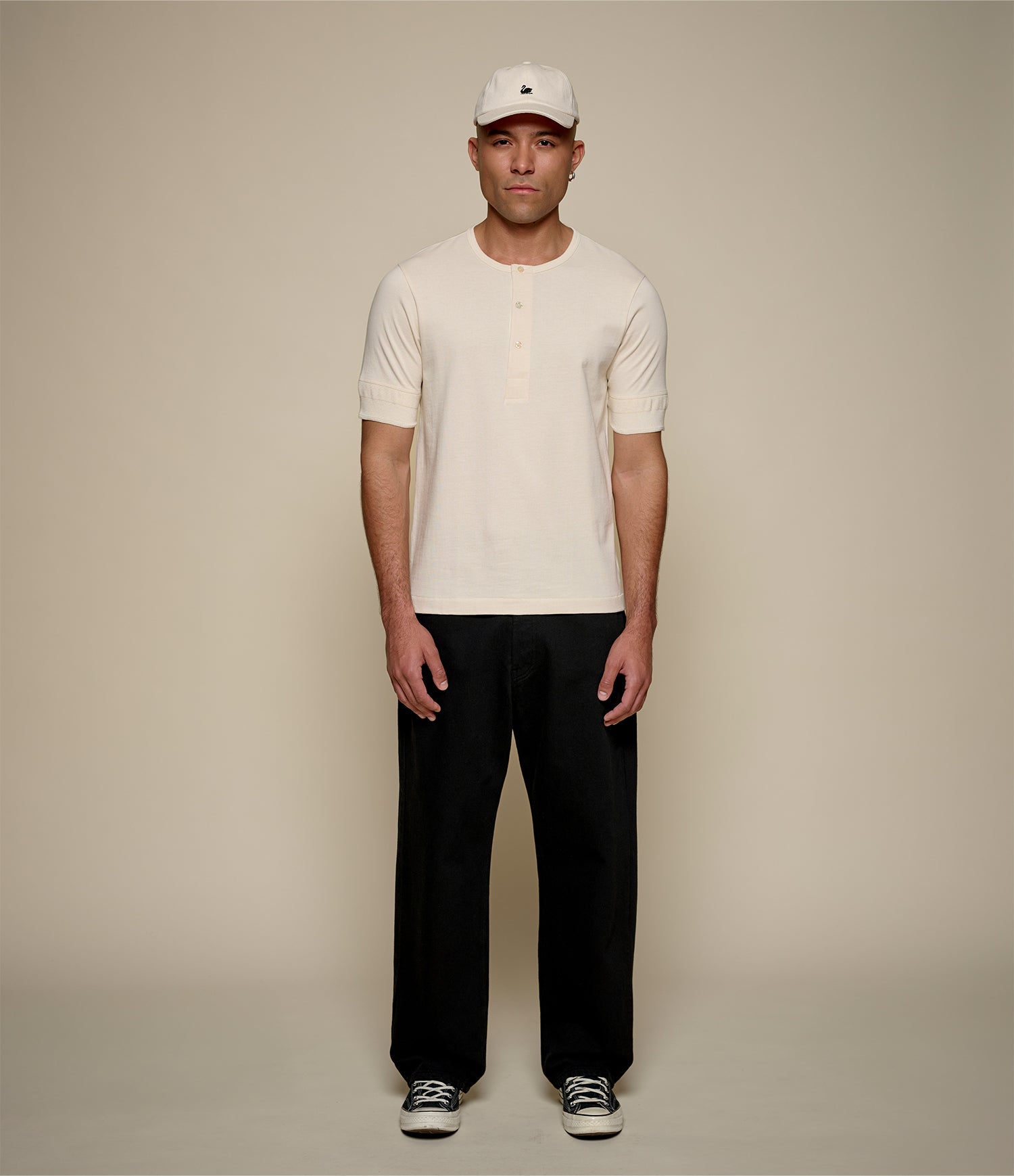 Man wearing a white cap, beige henley shirt, and black pants on a beige background