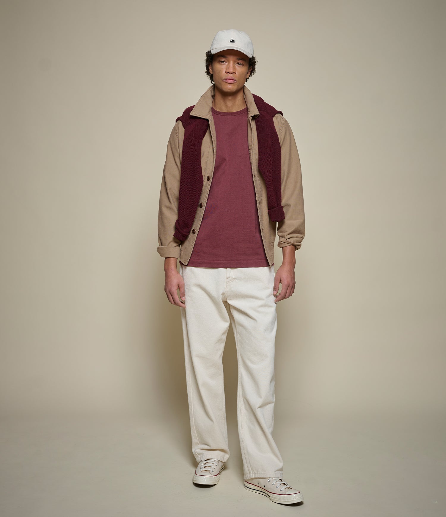 Man wearing a beige jacket with a maroon t-shirt, maroon shirt, white pants, and a white cap on a beige background