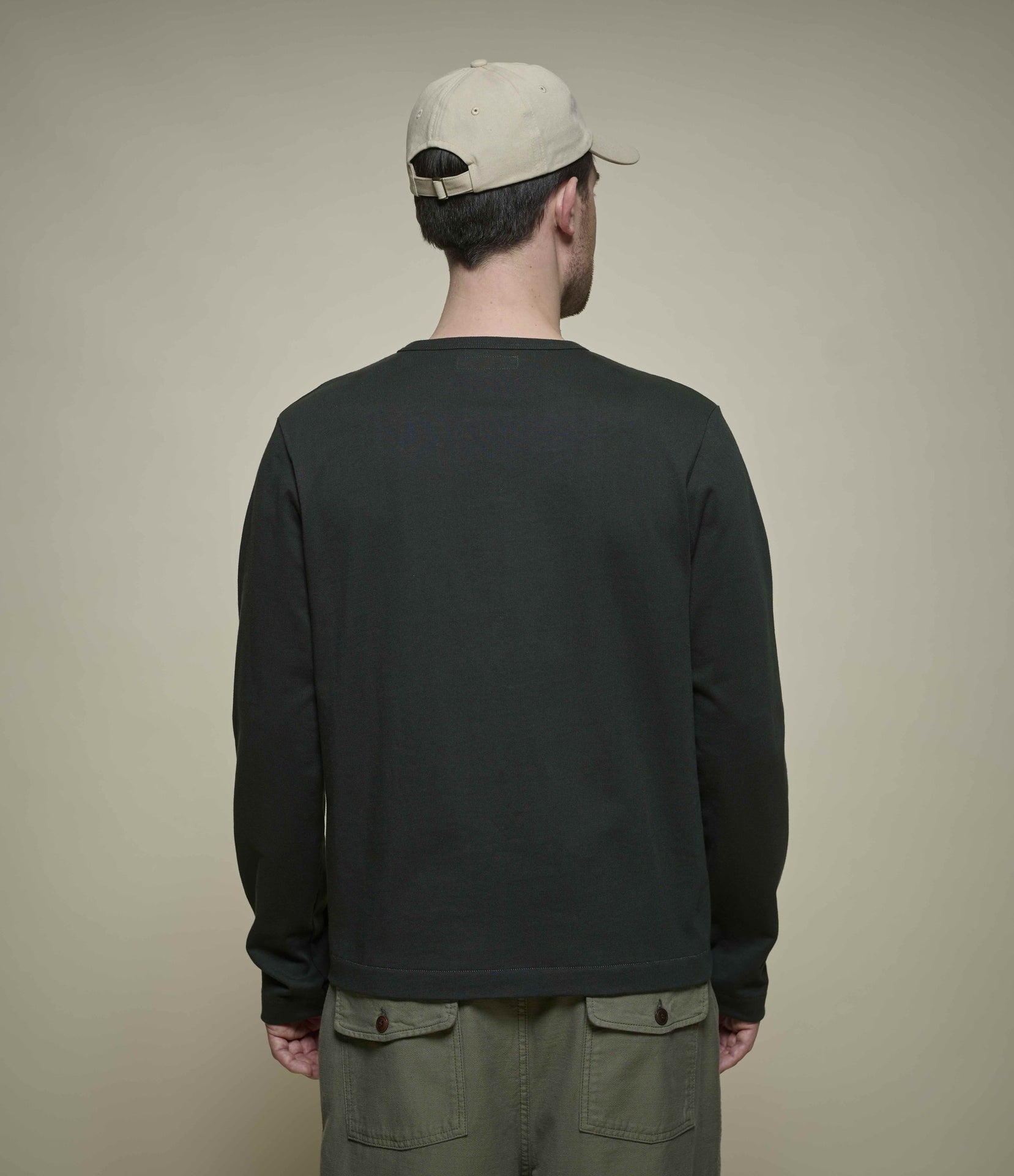 A man viewed from behind wearing a dark cropped henley, olive green cargo pants, and a beige baseball cap against a plain beige background.