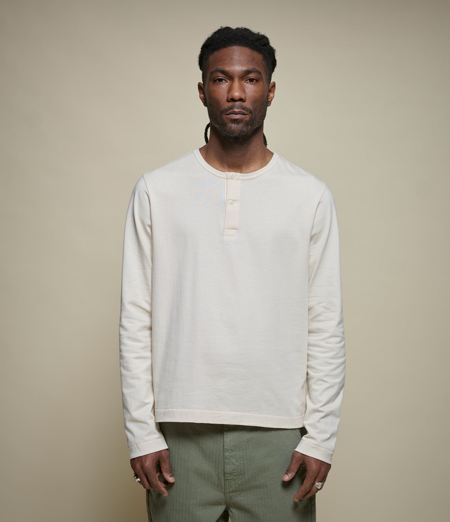 A man wearing a cream-colored cropped long-sleeve henley shirt and olive green pants, standing against a solid beige studio background.