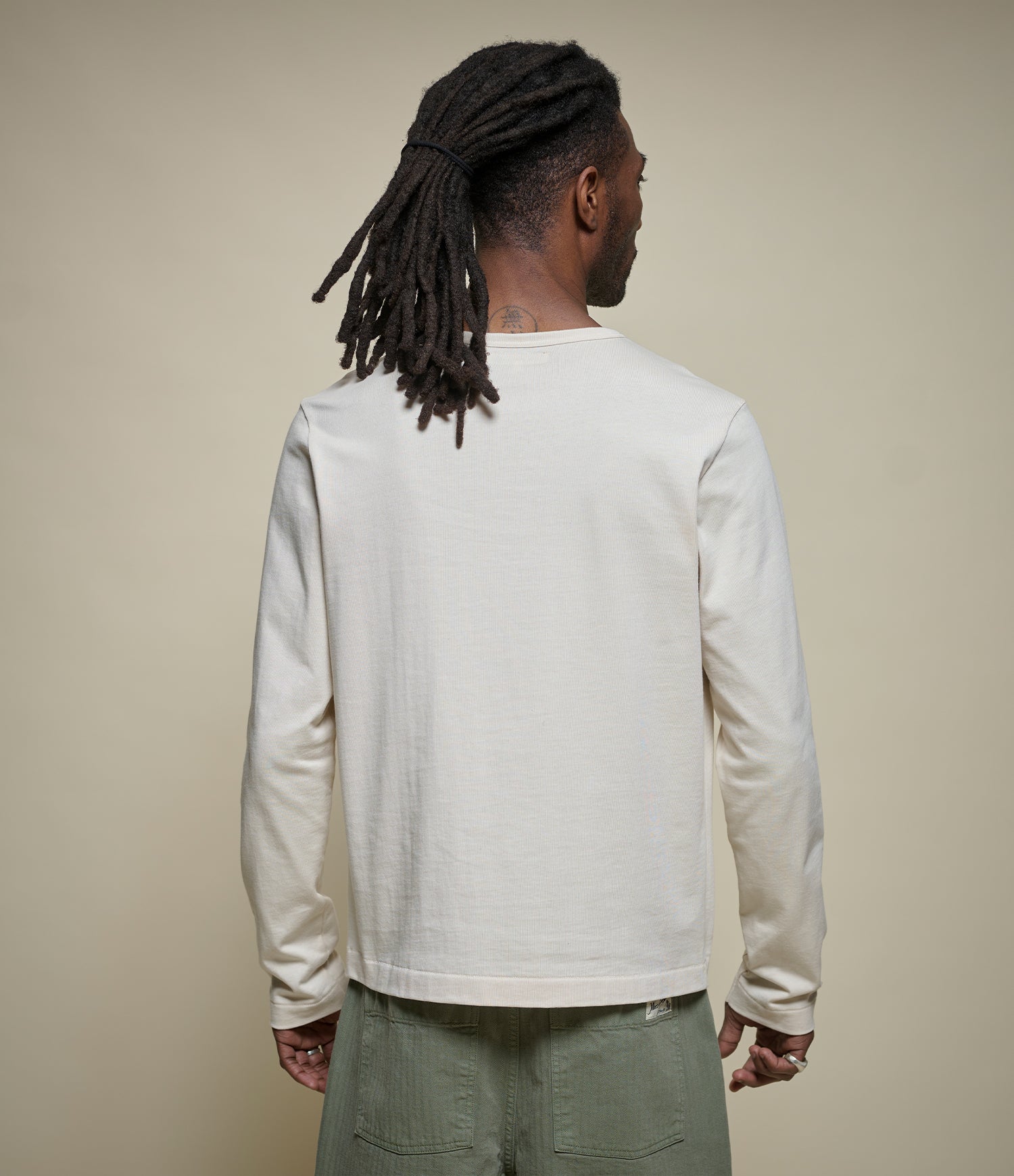 A man with long dark dreadlocks is shown from behind wearing a light cream/off-white long-sleeve henley and olive green pants, set against a plain beige studio background.