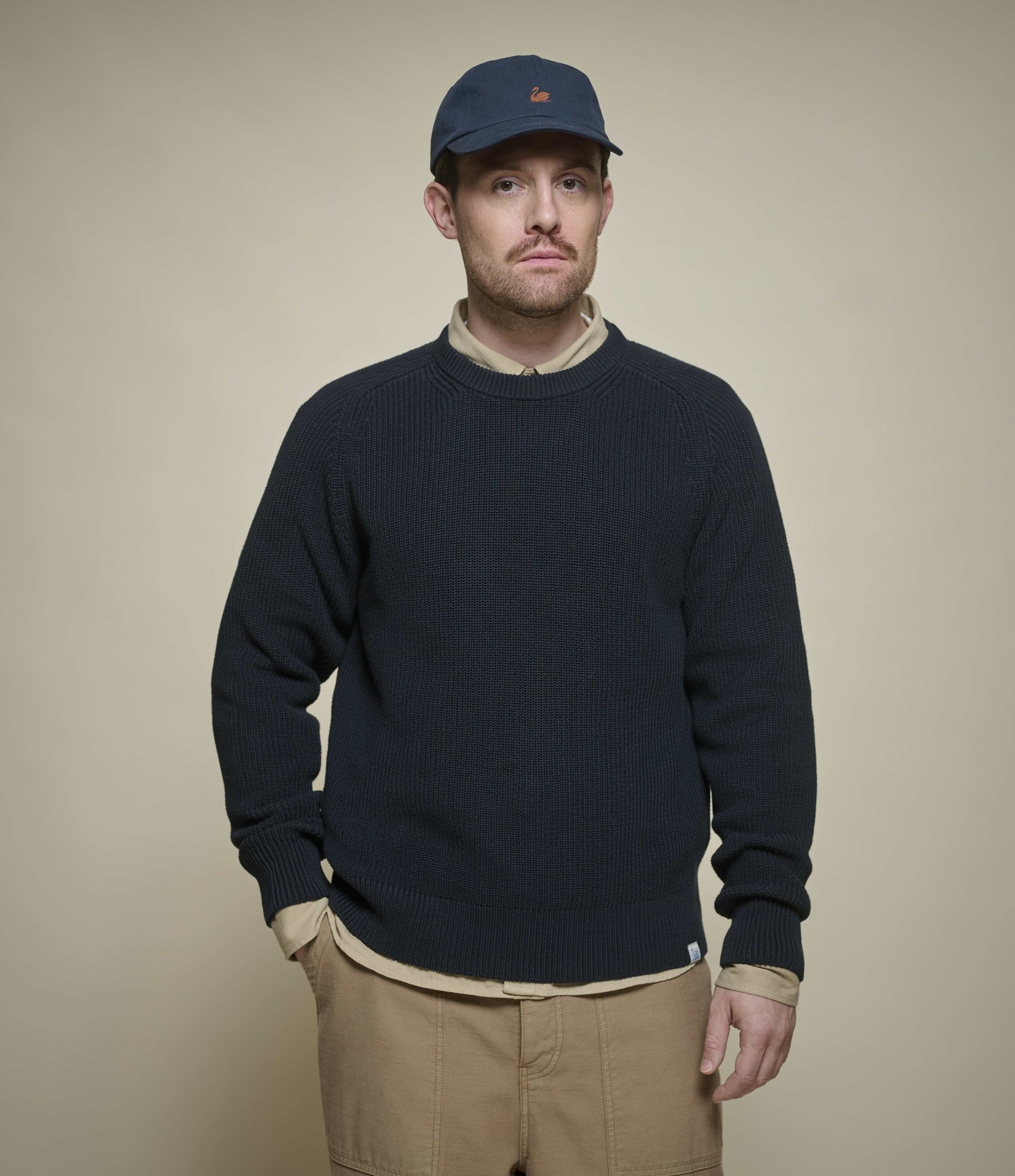 Man wearing a navy sweater, beige shirt, and cap against a beige background