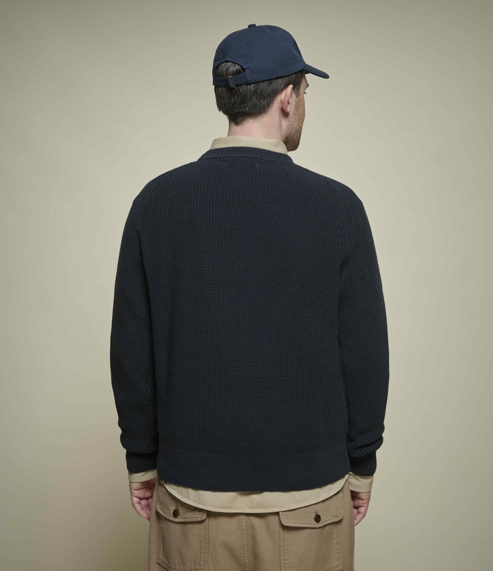 Man wearing a navy sweater and cap from behind on a beige background