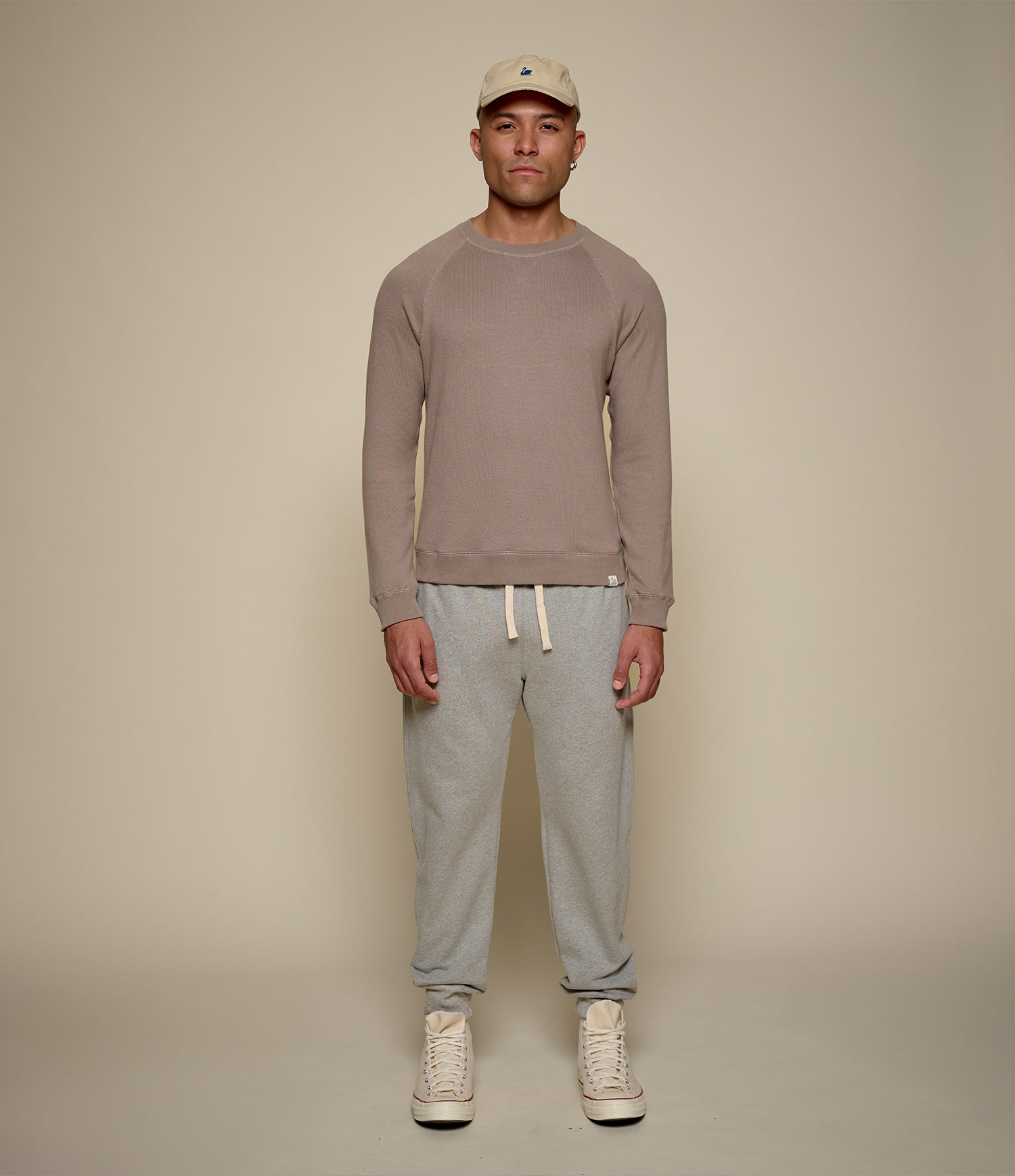 Man wearing a brown sweatshirt, gray sweatpants, and beige cap on a beige background