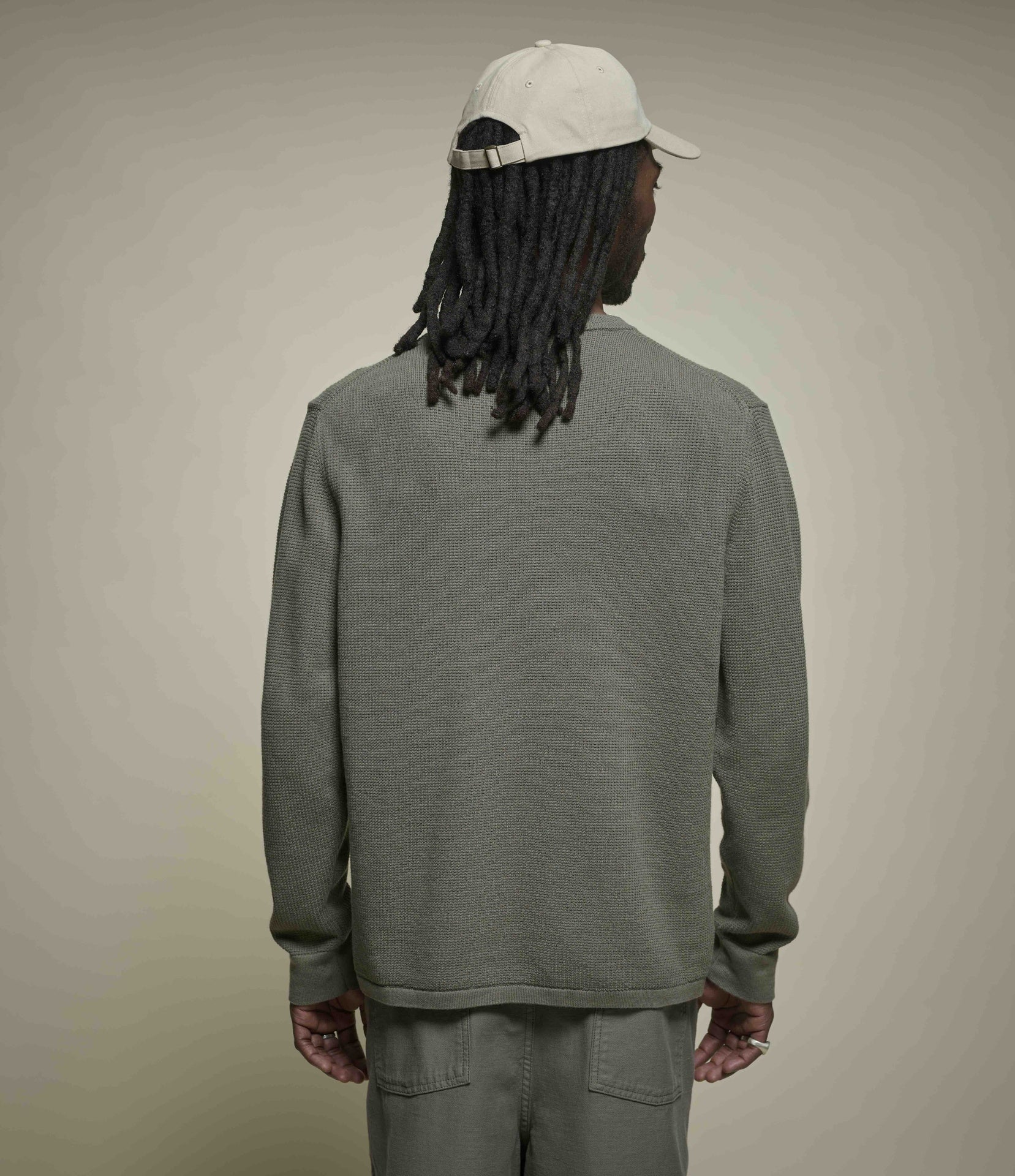 Person wearing an army green lightweight sweater and pants with a beige cap on a beige background