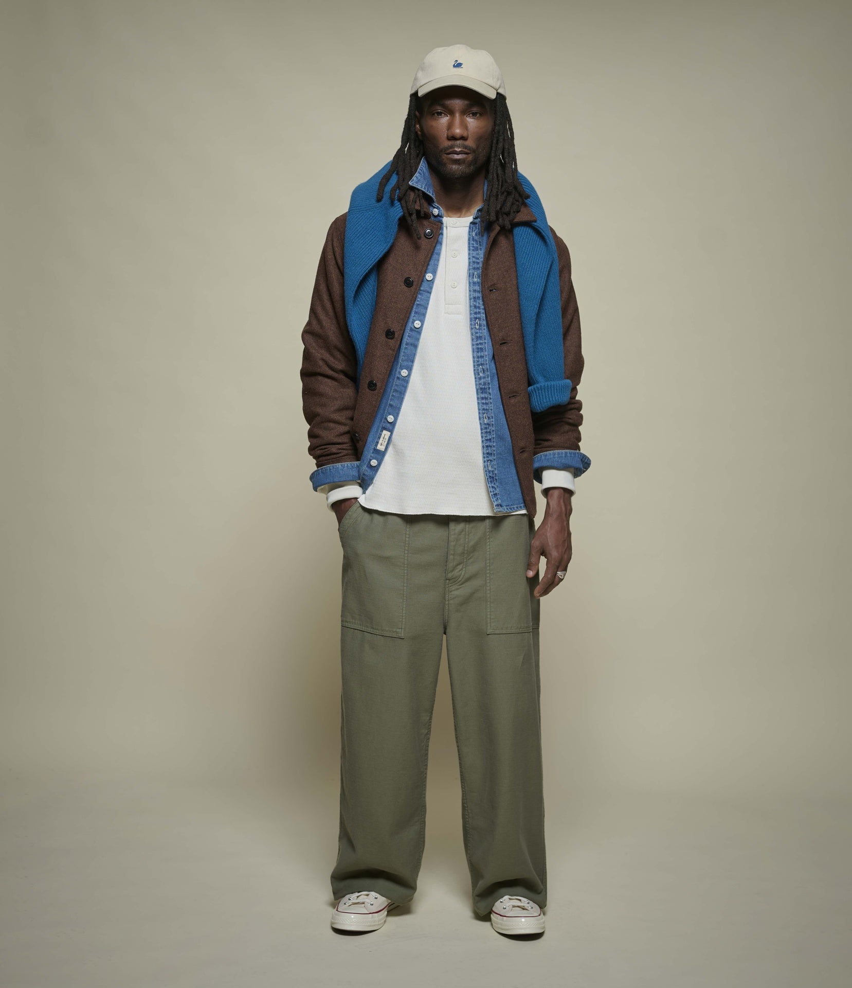 Person wearing a layered outfit with a brown jacket, blue shirt, beige henley shirt, green pants, and white sneakers on a beige background.