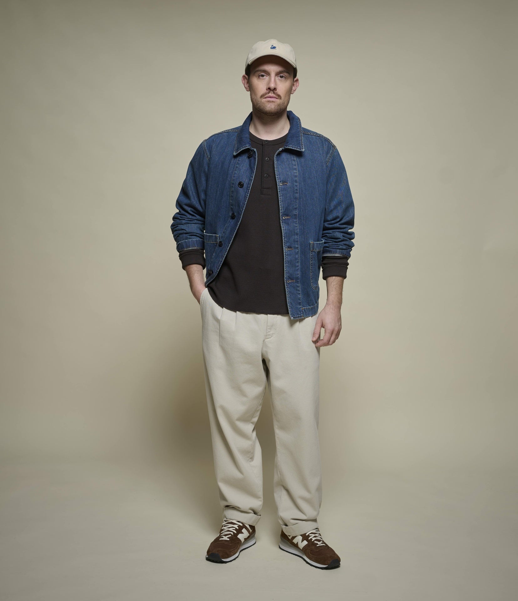 Man wearing a denim jacket, black shirt, beige pants, and brown shoes against a beige background