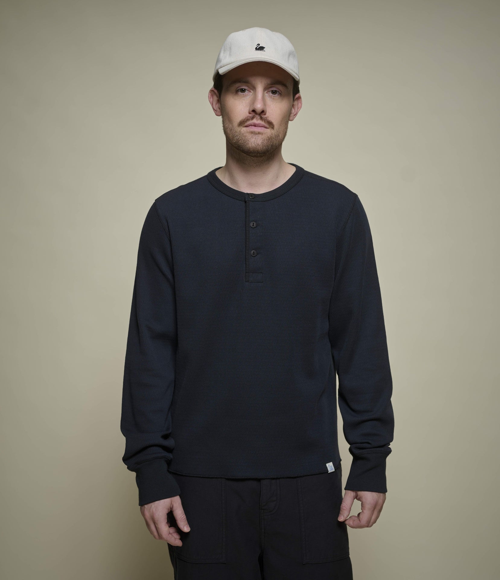Man wearing a black long-sleeve thermal henley and beige cap against a beige background