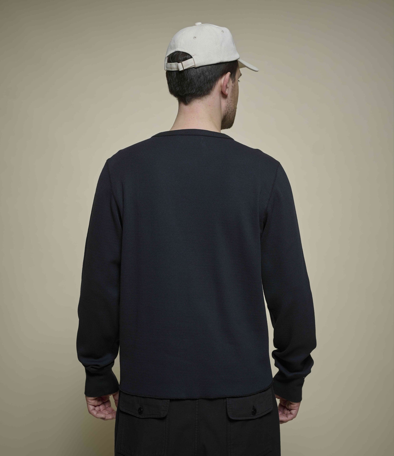 Man wearing a black long-sleeve henley shirt and white cap from behind on a beige background