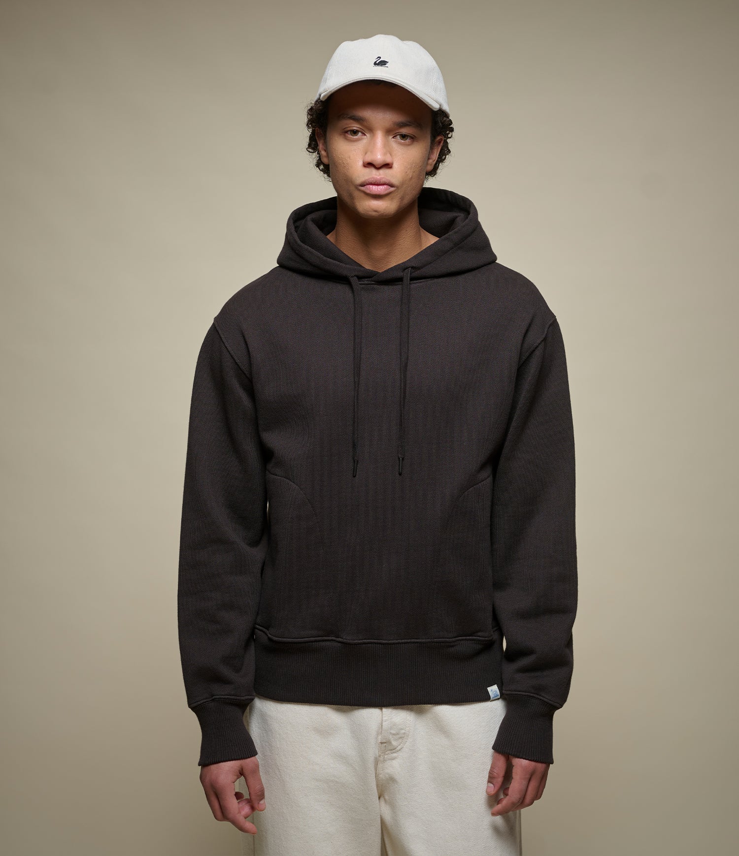 A model wearing a dark black ribbed pullover hoodie with black drawstrings, paired with off-white pants and a white baseball cap, standing against a solid beige studio background.