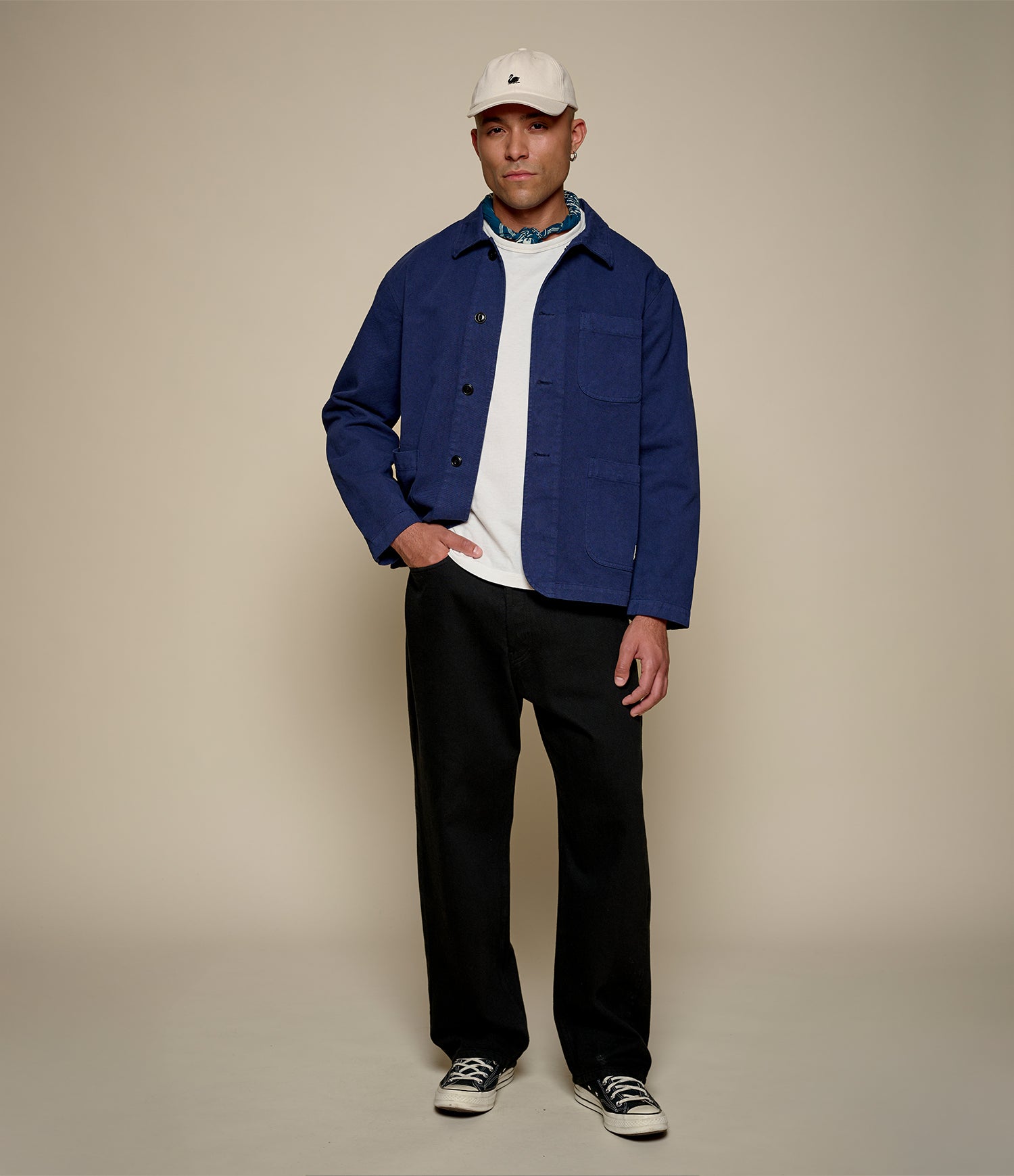 Man wearing a blue twill jacket, white shirt, black pants, and a beige cap on a beige background