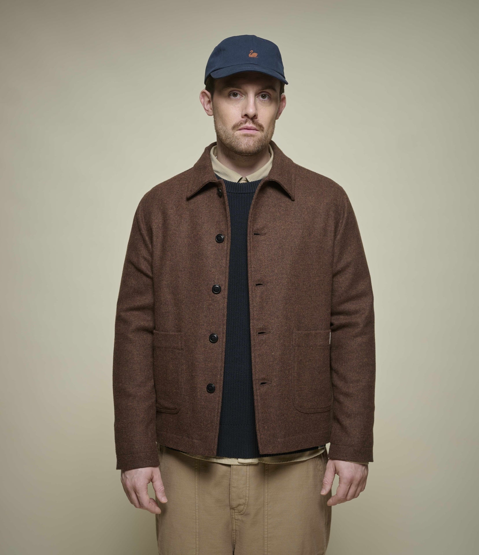 A man wearing a brown wool tweed button-front jacket over a dark shirt, khaki pants, and a navy baseball cap, standing against a plain beige studio background.