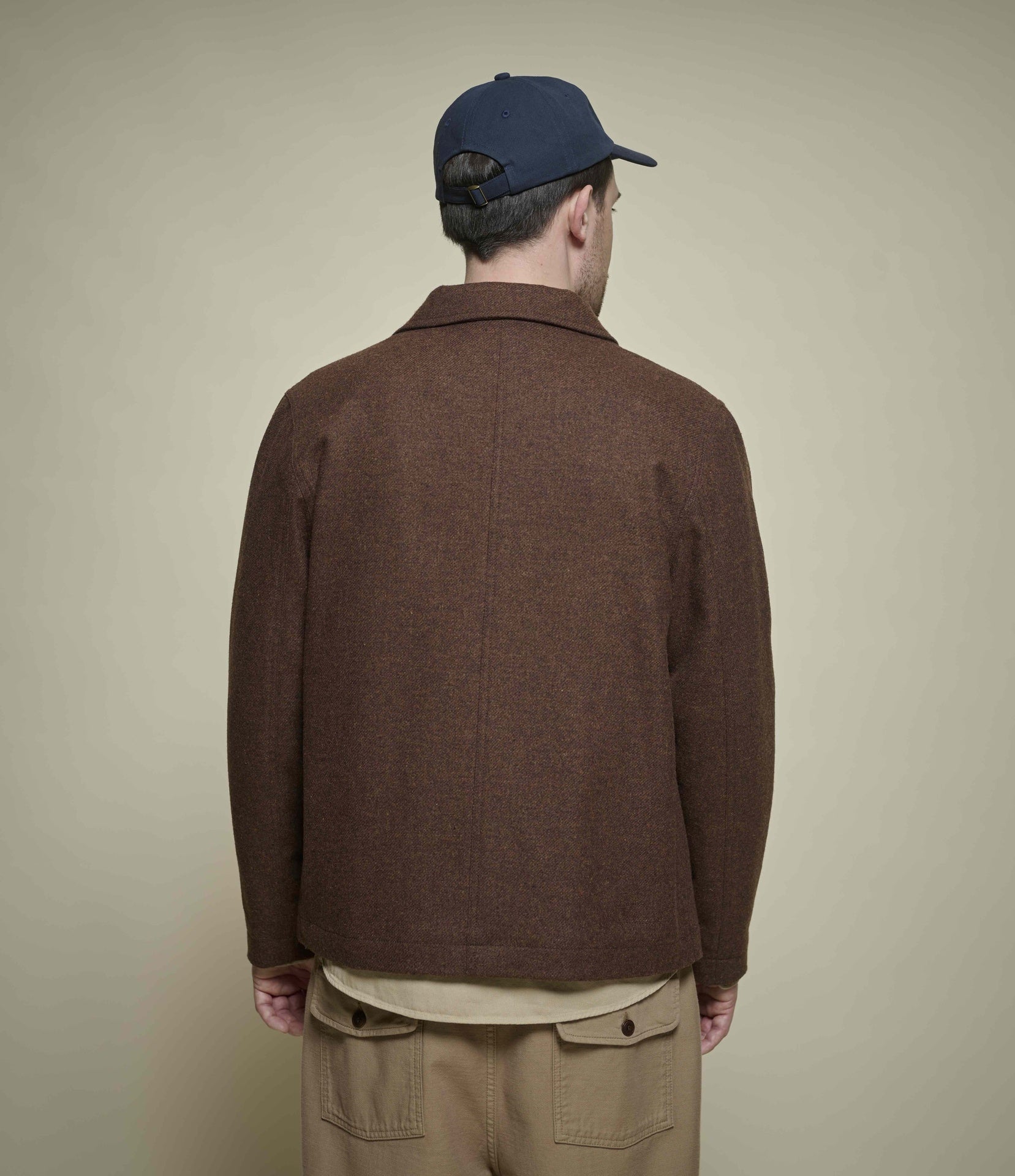 A man viewed from behind wearing a brown wool tweed jacket, khaki pants, and a navy baseball cap against a plain beige studio background.