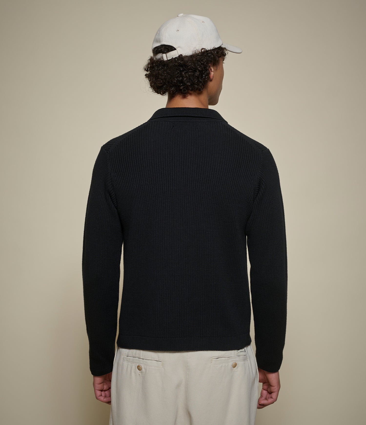 A man shown from the back wearing a black ribbed knit long-sleeve polo shirt, cream pants, and a white baseball cap against a plain beige background.
