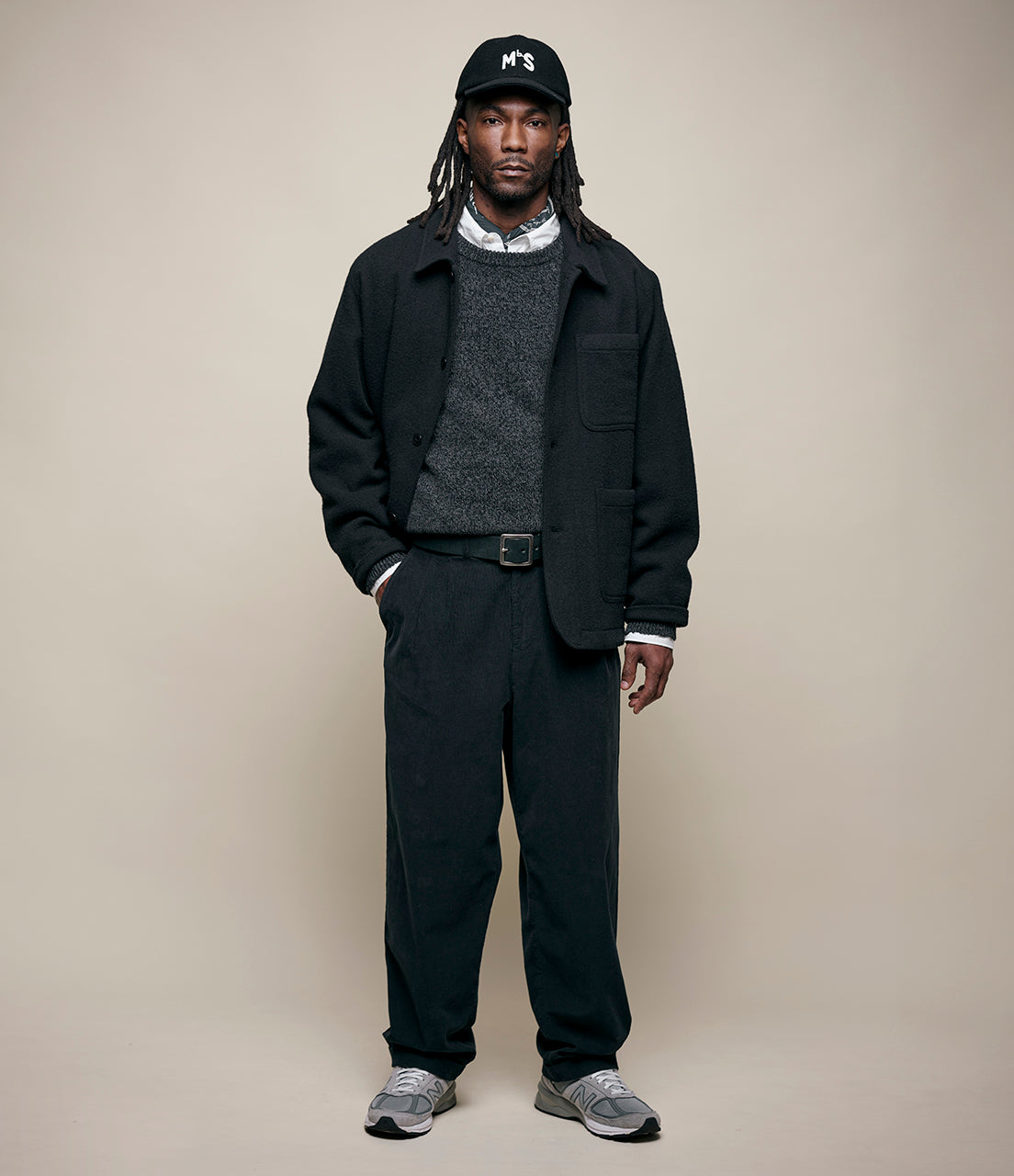 Man wearing a black jacket, gray sweater, and pants with a cap on a beige background