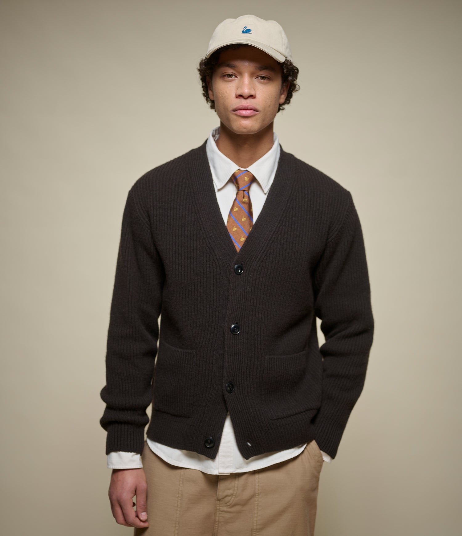 A man wearing a dark brown ribbed knit cardigan over a white collared shirt with an orange patterned tie, khaki pants, and a beige baseball cap, standing against a solid beige background.