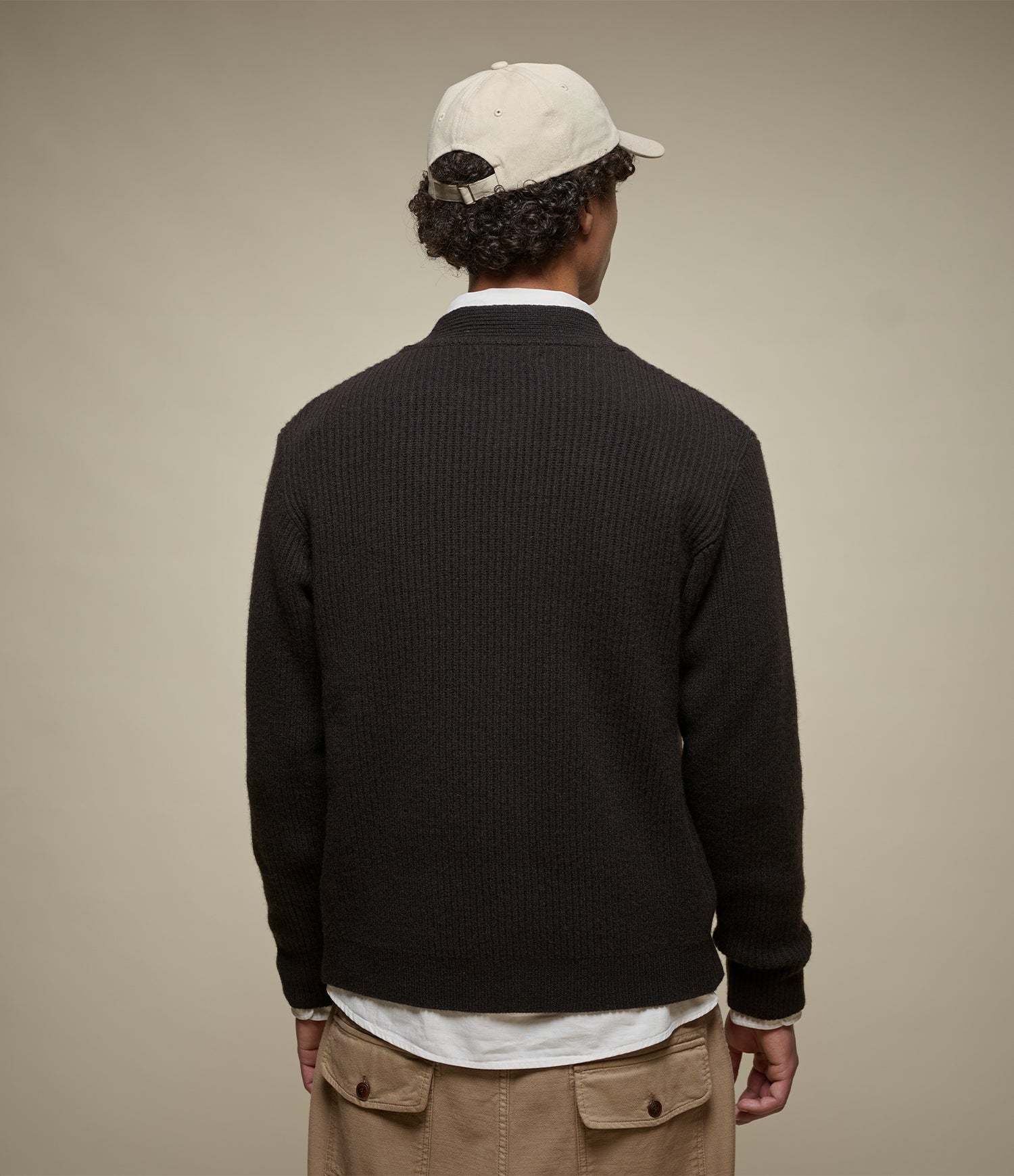 A man viewed from behind wearing a dark brown ribbed knit cardigan over a white shirt, khaki pants, and a cream baseball cap, against a solid beige background.
