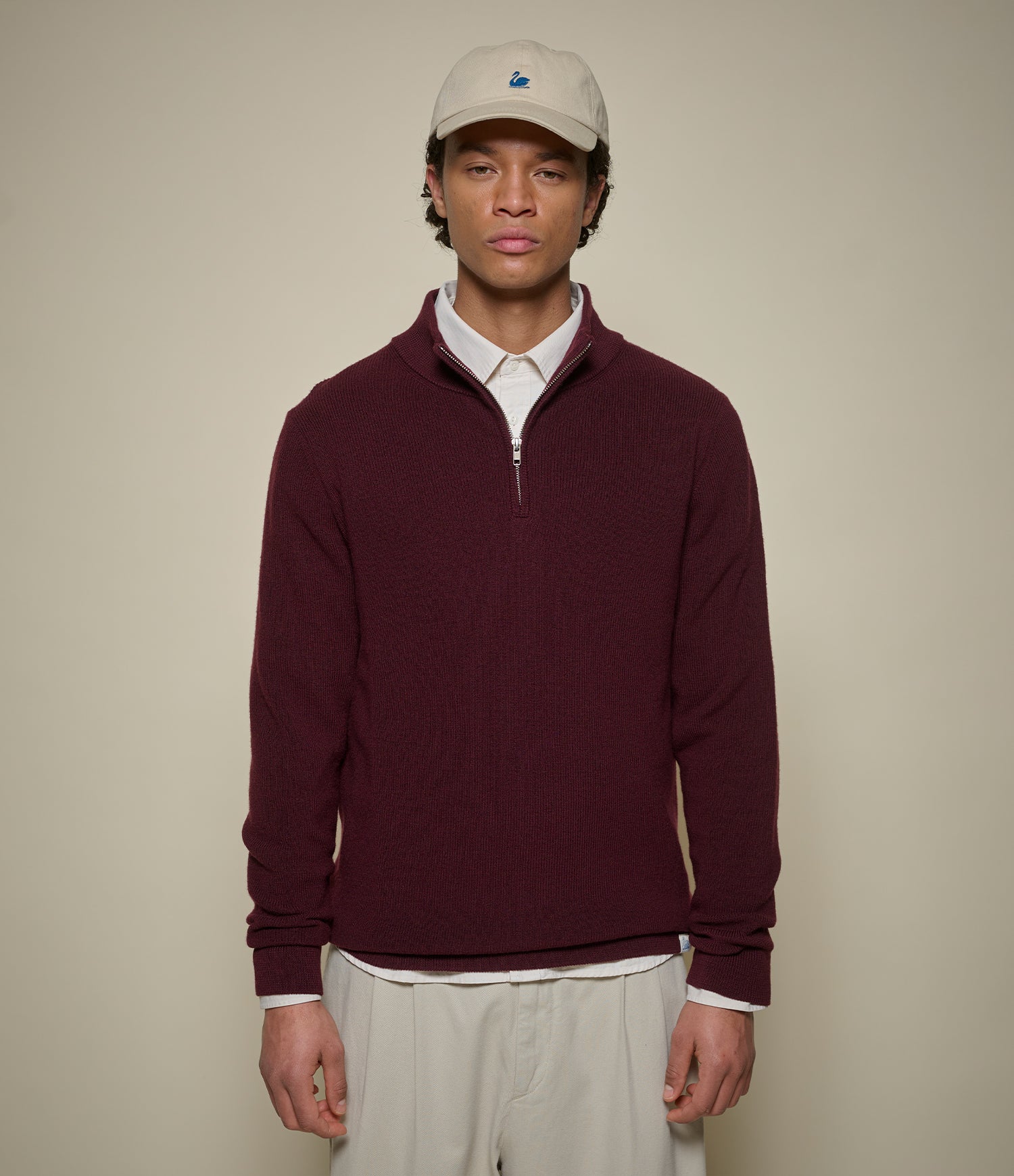 Man wearing a burgundy half-zip pullover with a white cap against a beige background