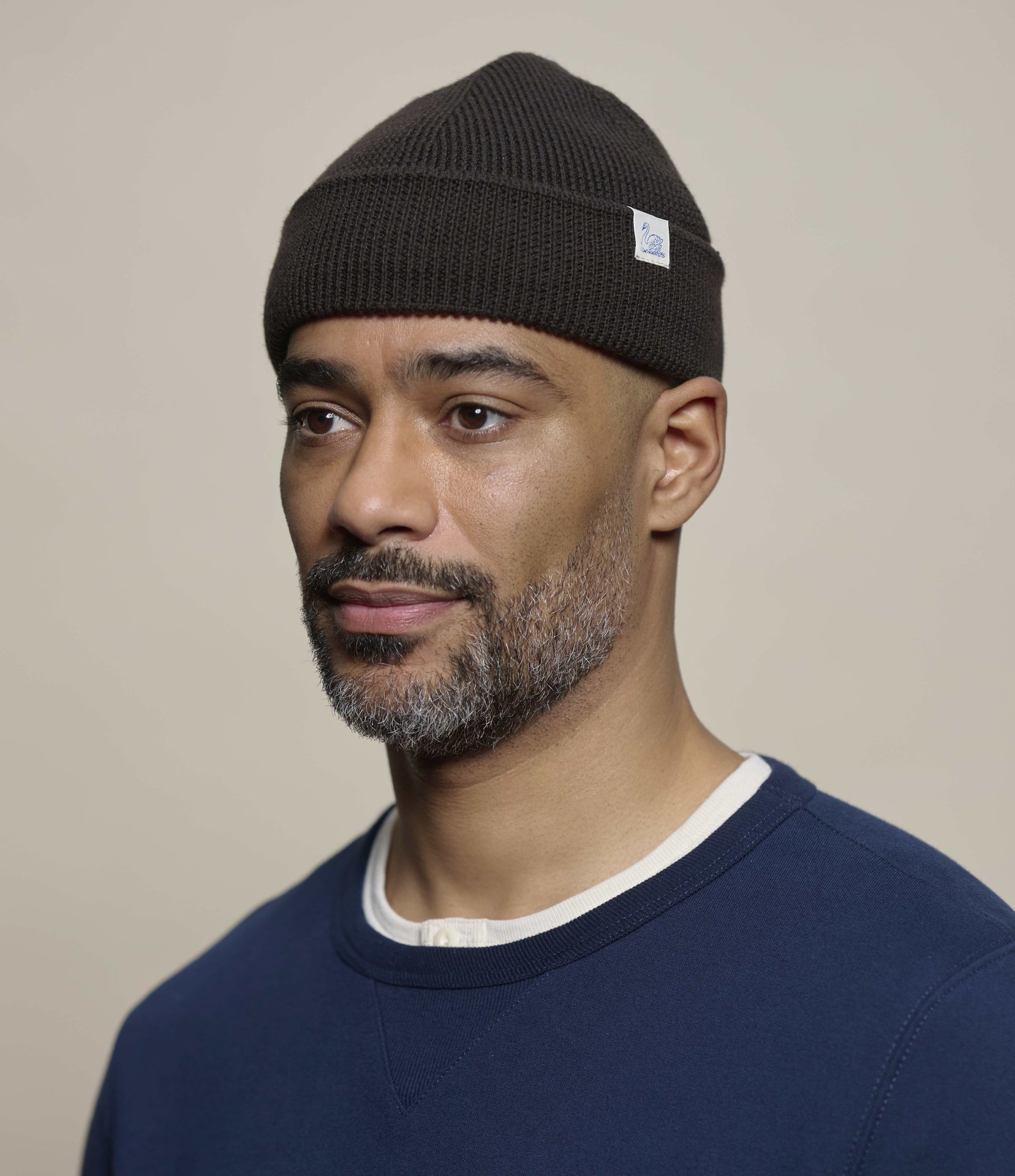 A man wearing a dark brown ribbed knit beanie with a small white brand tag on the side, paired with a navy blue crewneck sweater. The background is a solid, warm beige studio backdrop.