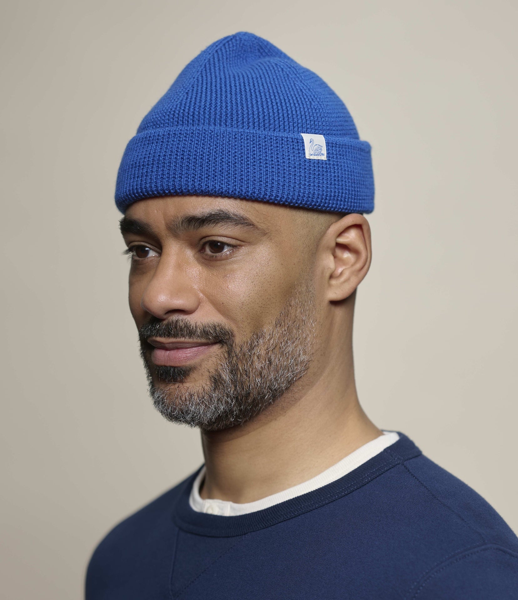 A man wearing a bright blue knit beanie with a small white square logo patch on the cuff, photographed against a plain warm beige studio background.