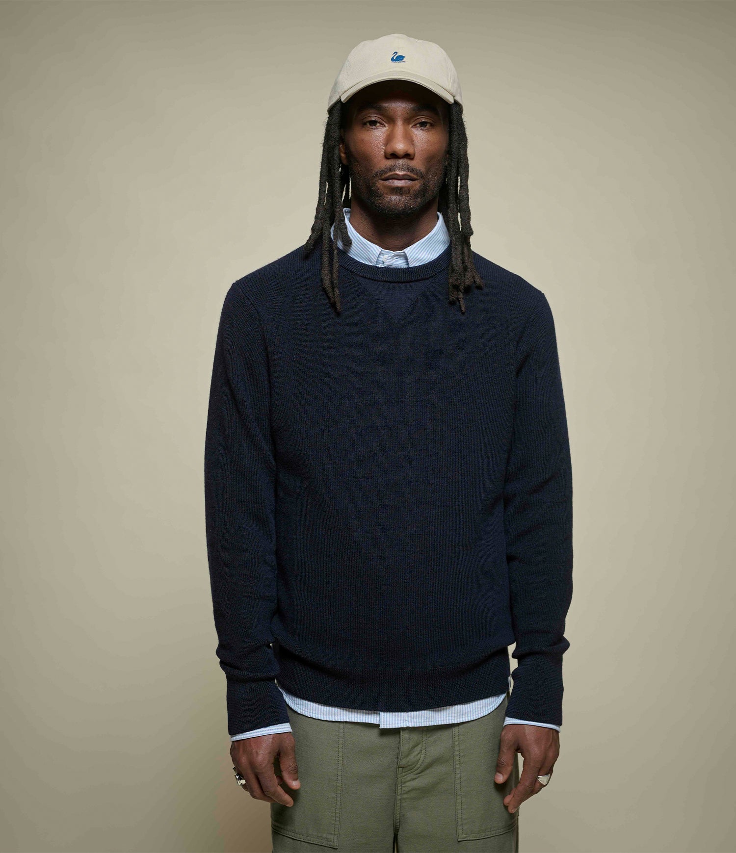 Man wearing a navy merino wool sweater, beige cap, and light-colored pants against a beige background