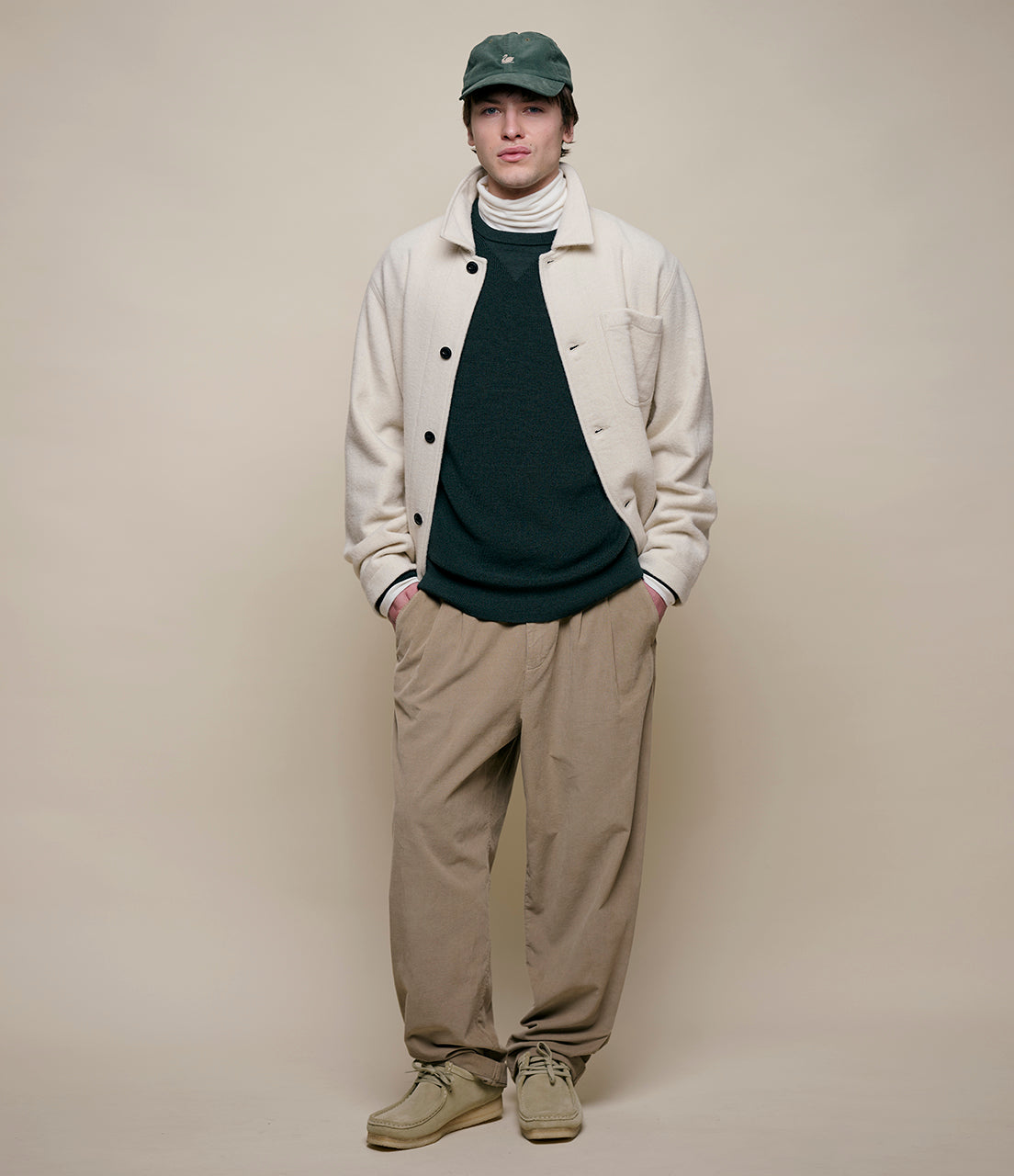 Person wearing a beige jacket, dark shirt, beige pants, and green cap against a plain background
