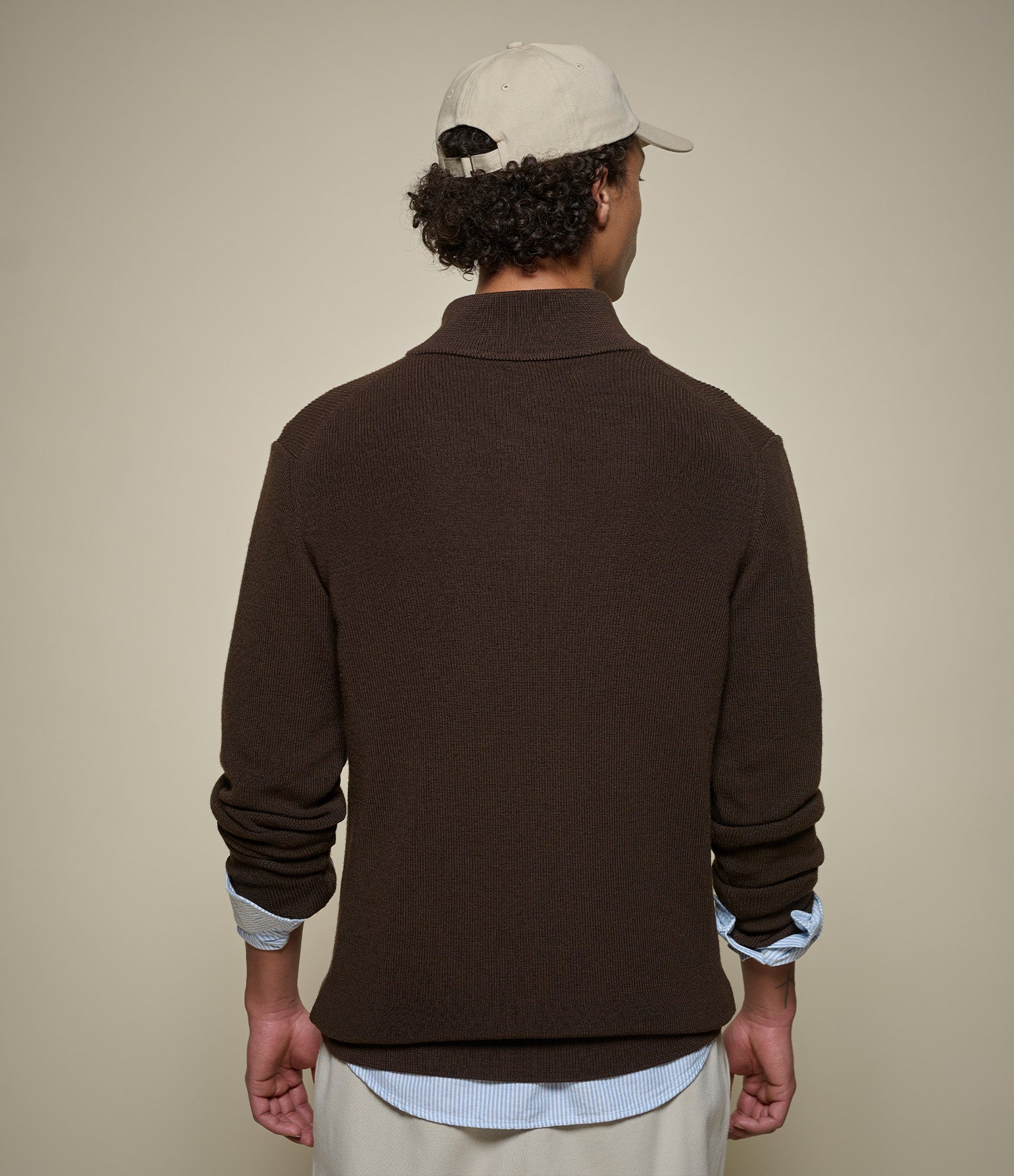 Man wearing a brown sweater over a light blue shirt with a beige cap on a beige background