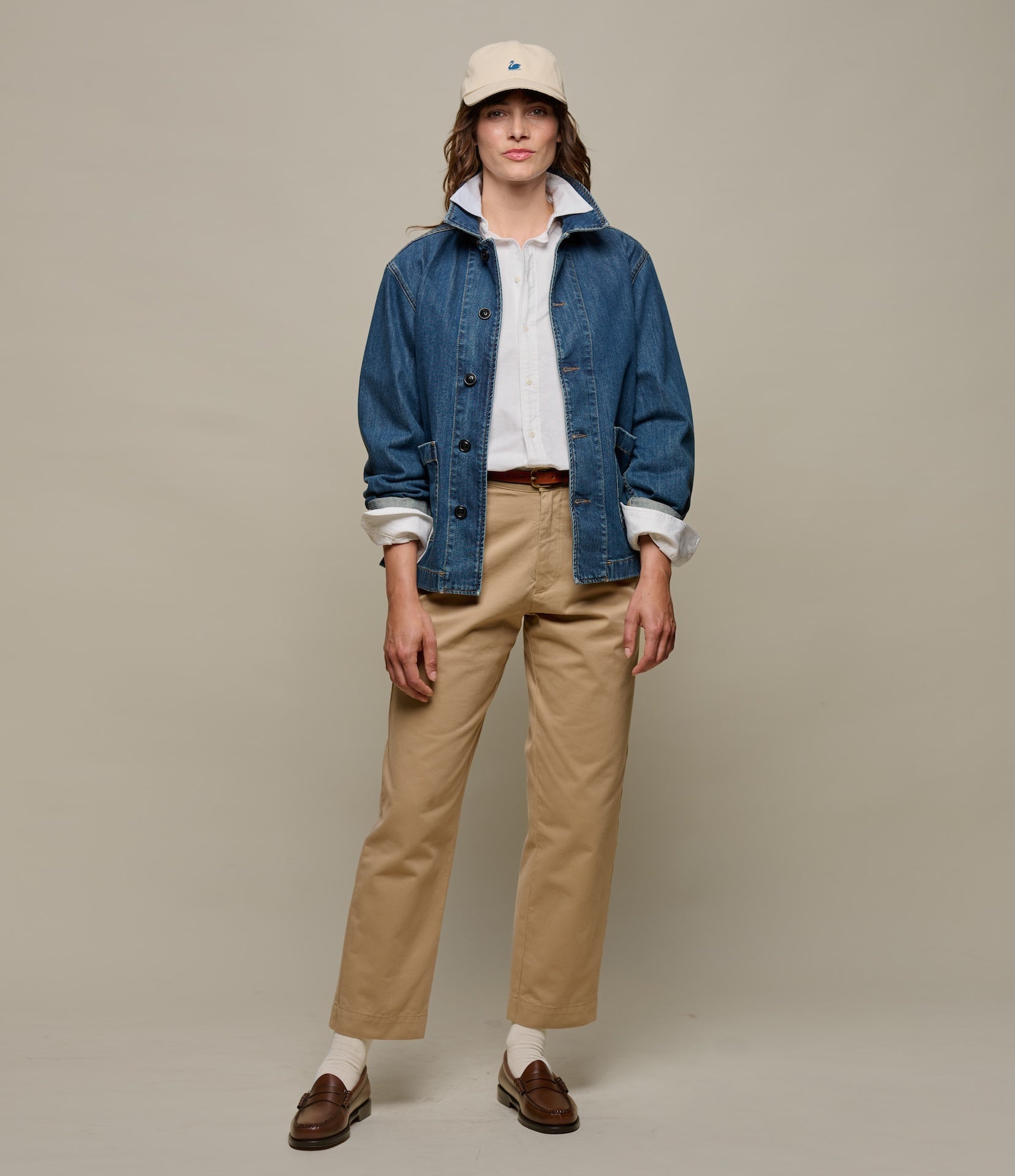 Woman wearing a blue denim jacket, beige pants, and a beige cap on a plain background