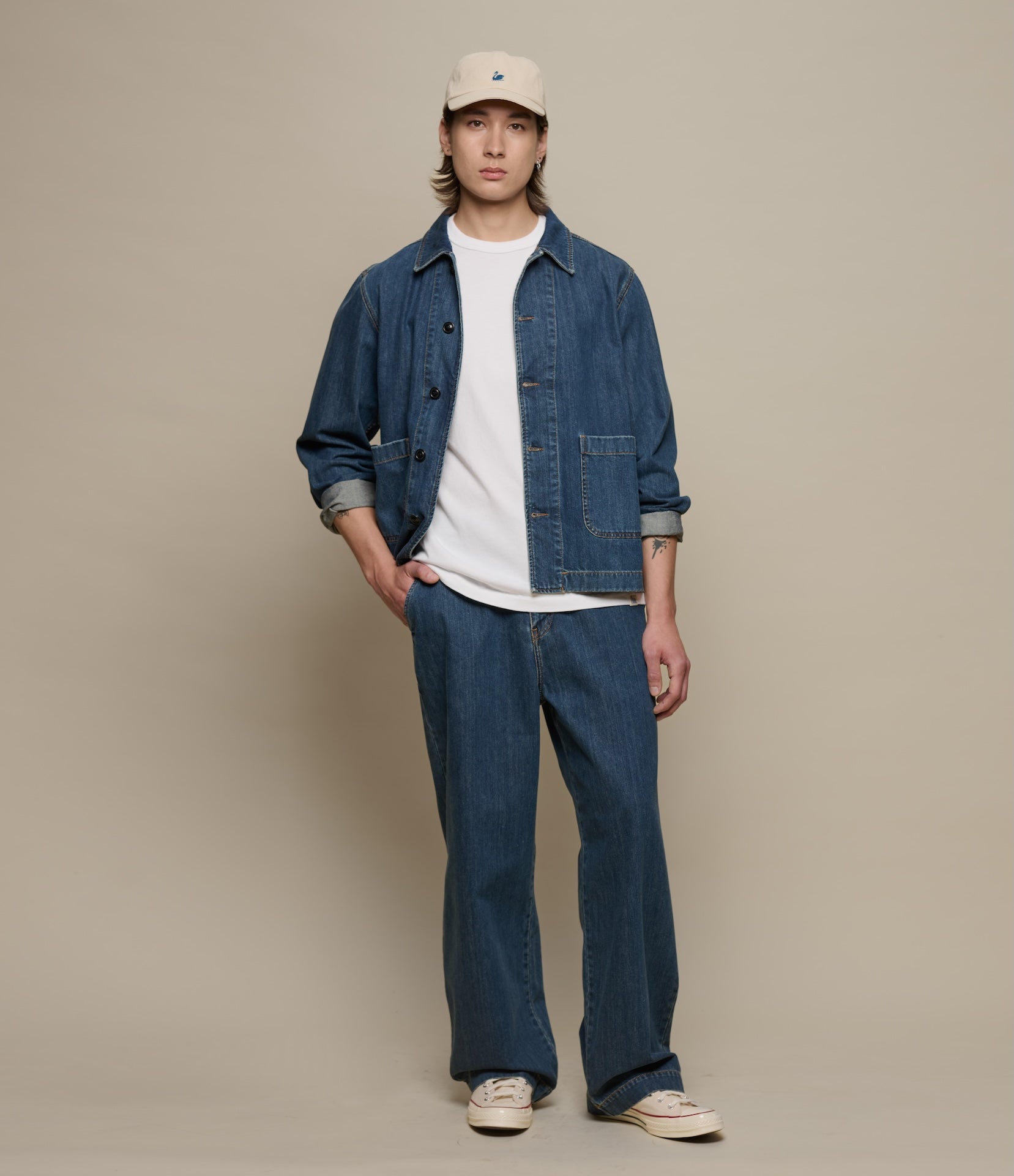 Person wearing a denim jacket, white shirt, and blue jeans against a beige background