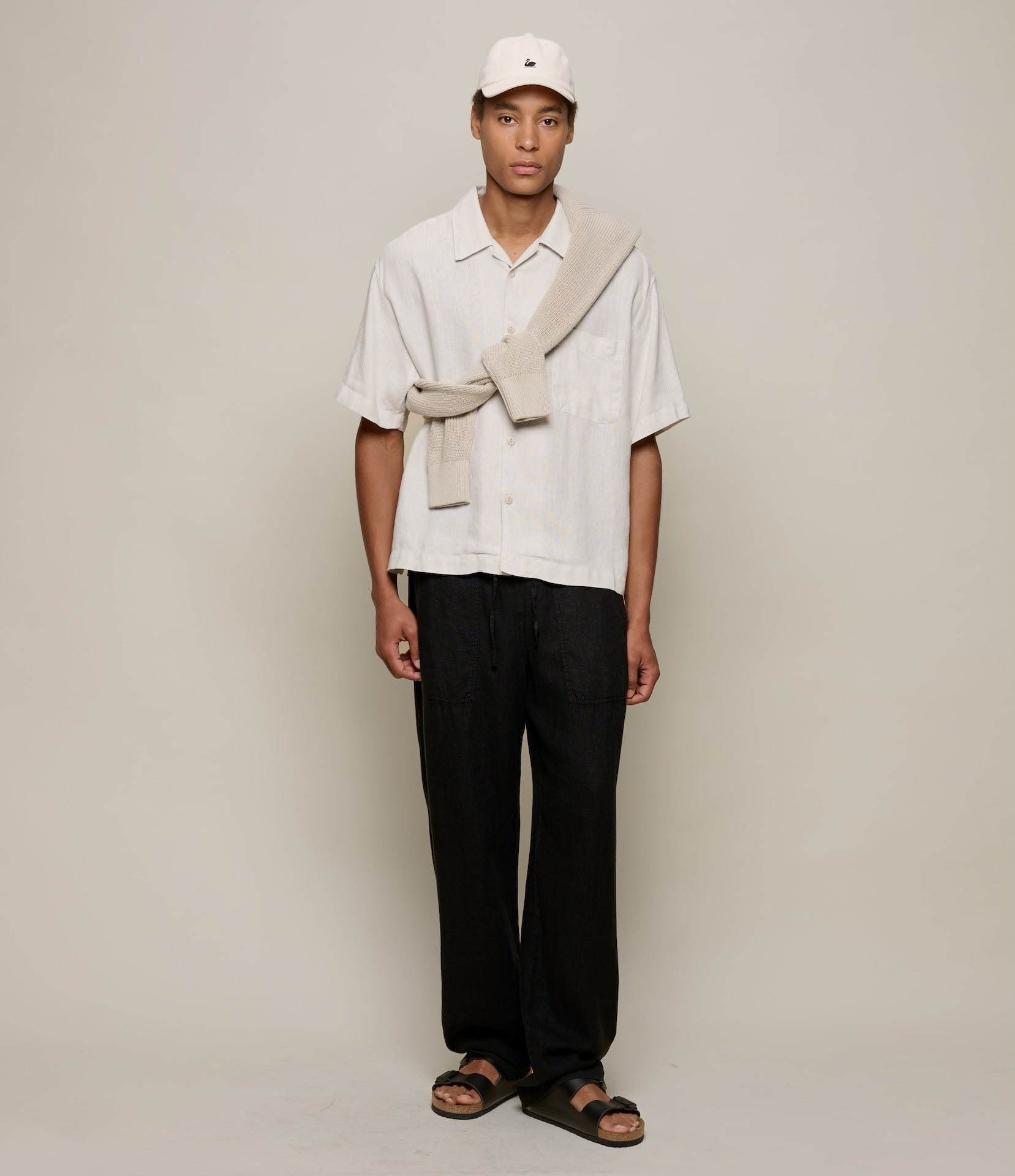 Person wearing a beige linen shirt, black pants, and a beige scarf against a plain background