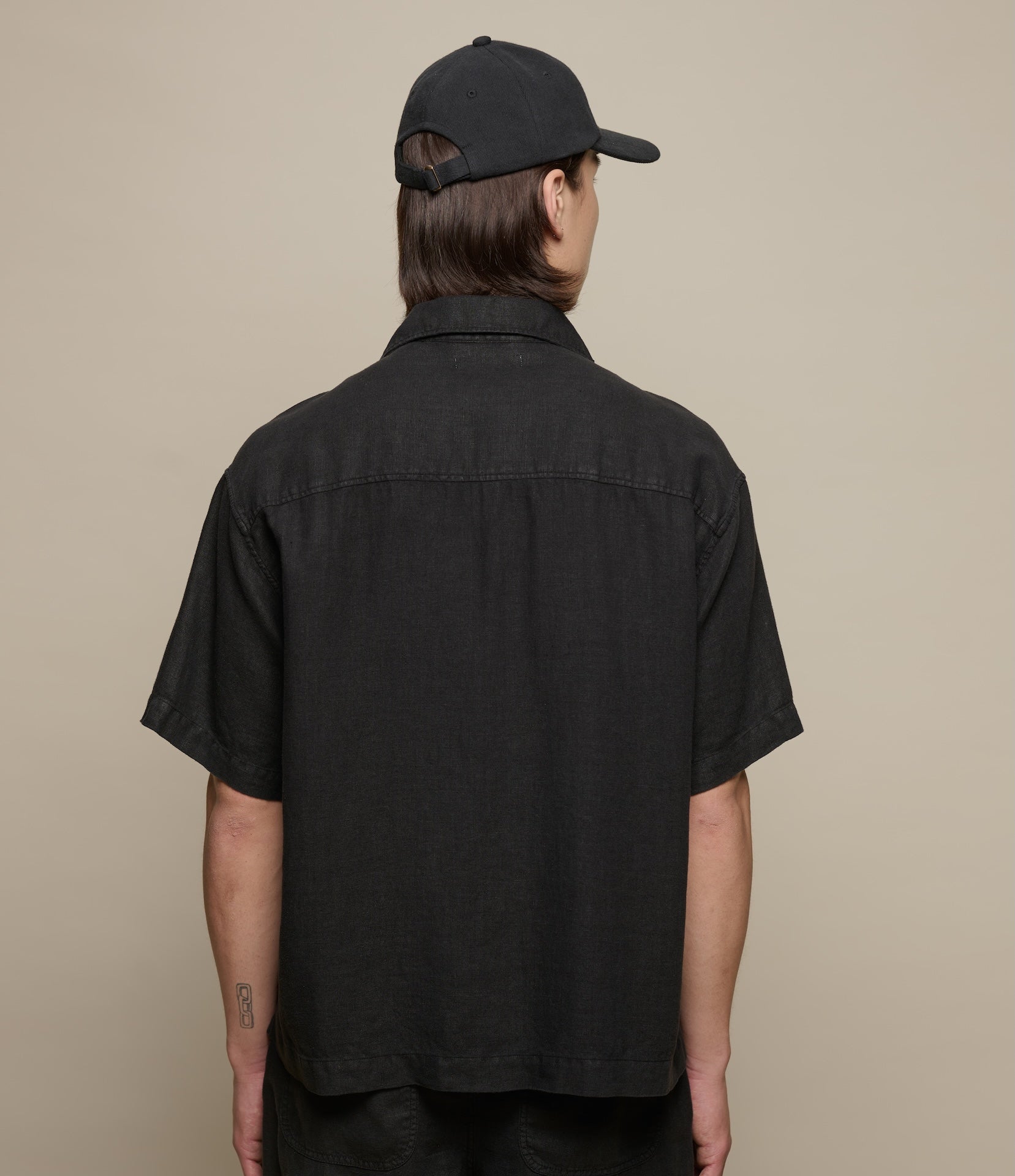 Person wearing a black short-sleeve linen shirt and cap from behind on a beige background