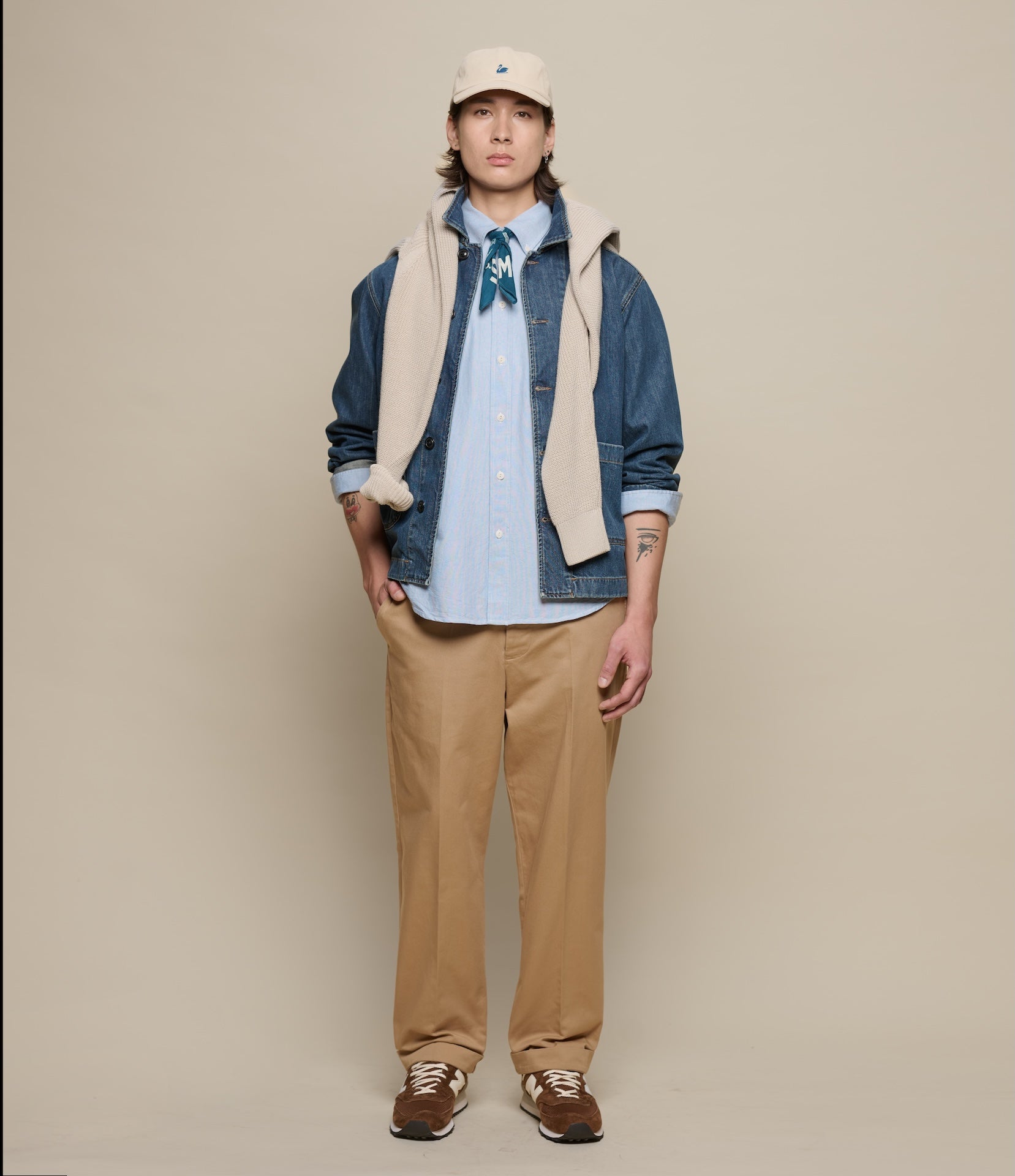 Person wearing a denim jacket, beige cap, and tan pants against a beige background