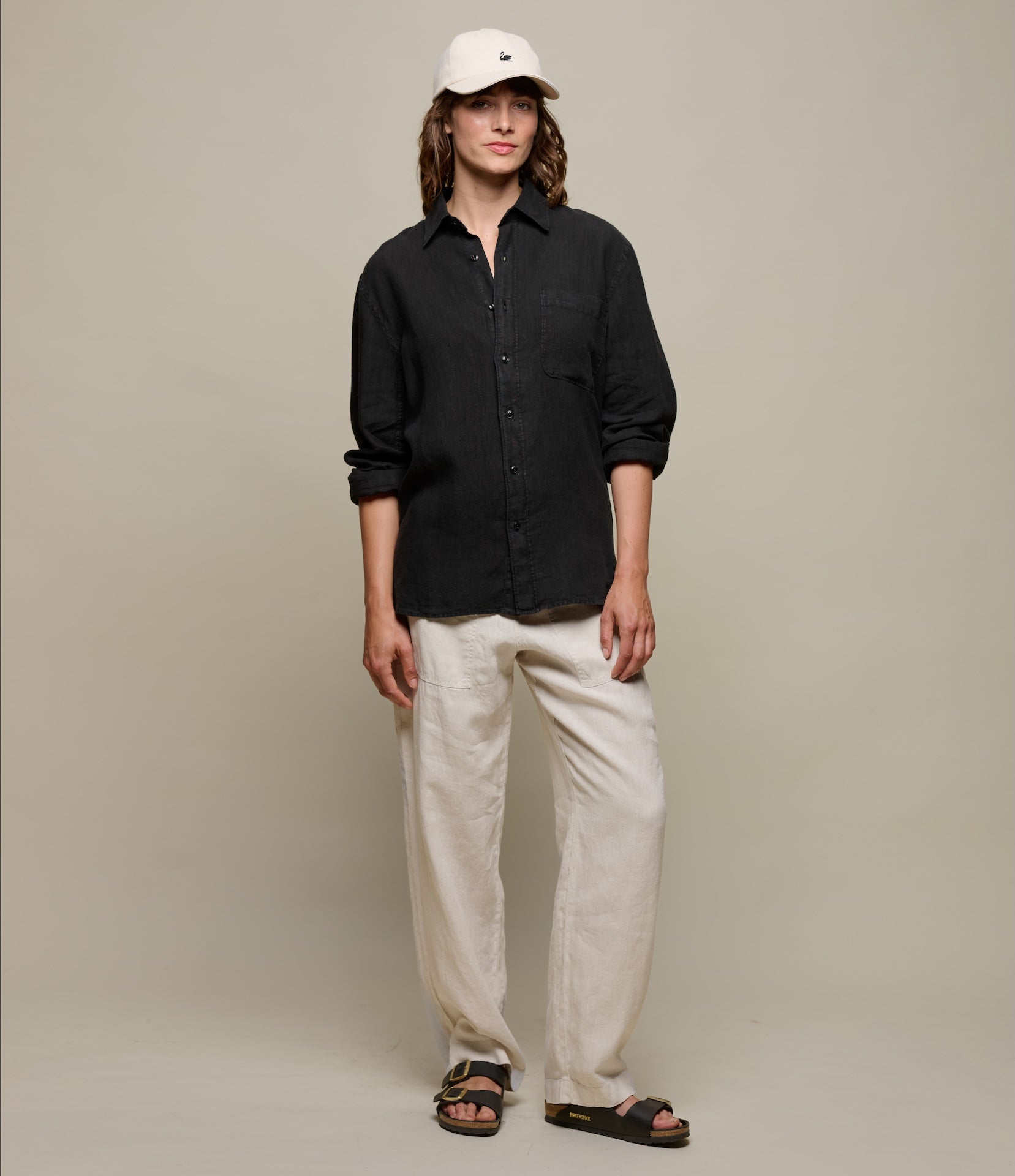 Person wearing a black linen shirt, white pants, and a white cap against a plain background