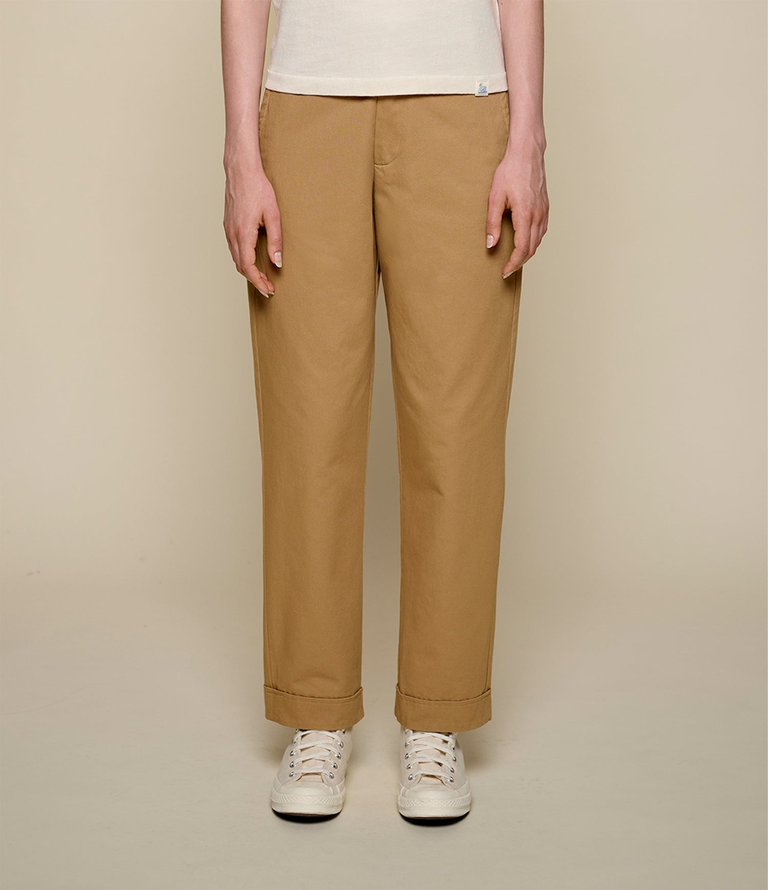 A person wearing straight-leg tan chinos with white sneakers, shown from the waist down against a solid beige studio background.