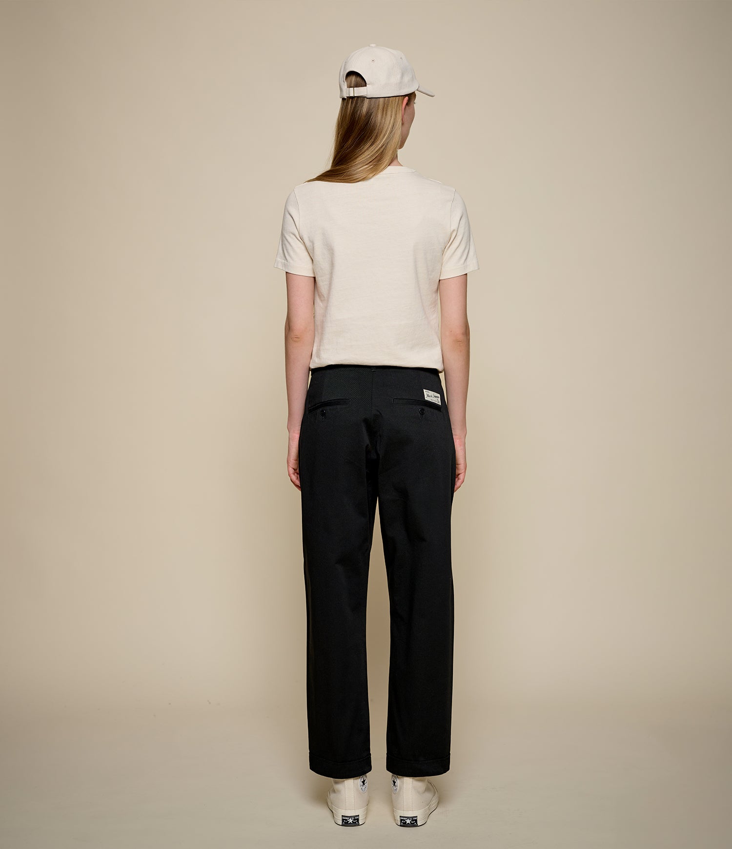 A woman is shown from the back wearing a cream short-sleeve t-shirt, black wide-leg trousers, a cream baseball cap, and white sneakers against a solid beige studio background.
