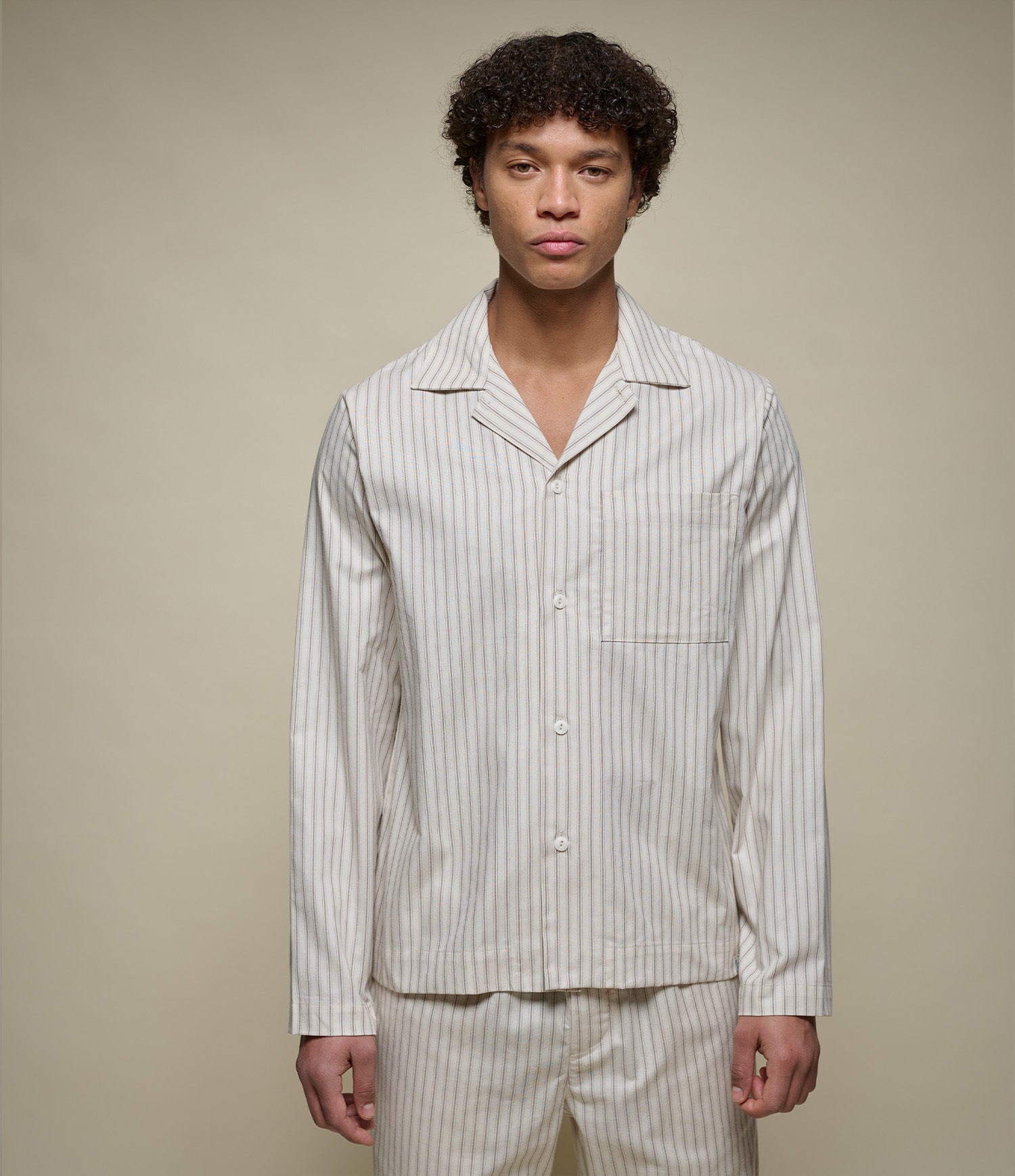 A man wearing a matching long-sleeve button-front pajama top and trousers set with thin vertical stripes, photographed against a plain warm beige studio background.
