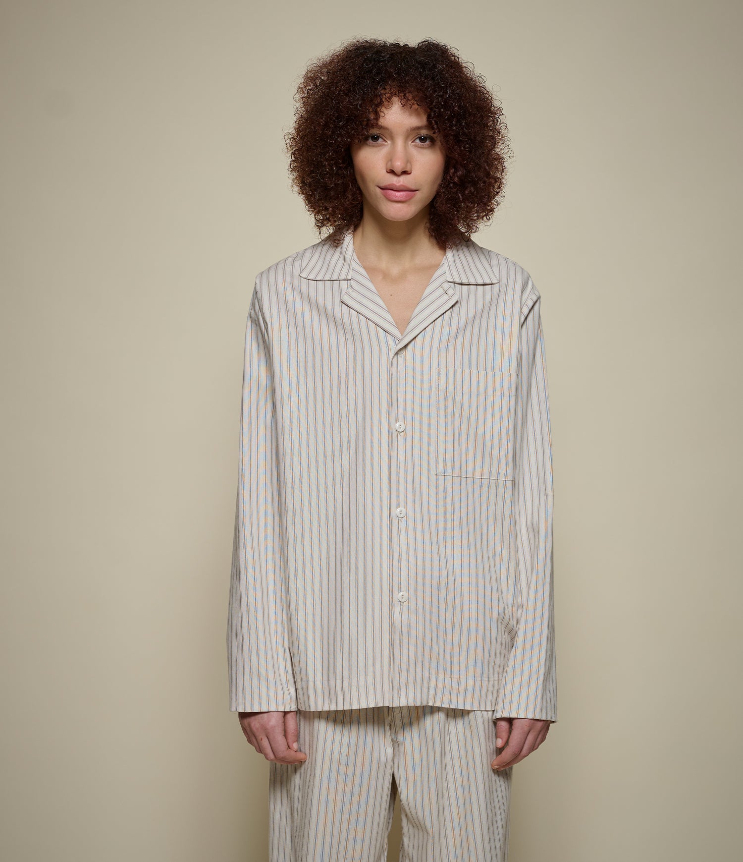 A model with curly brown hair wearing a matching two-piece pajama set with beige and white vertical stripes, standing against a solid beige background.