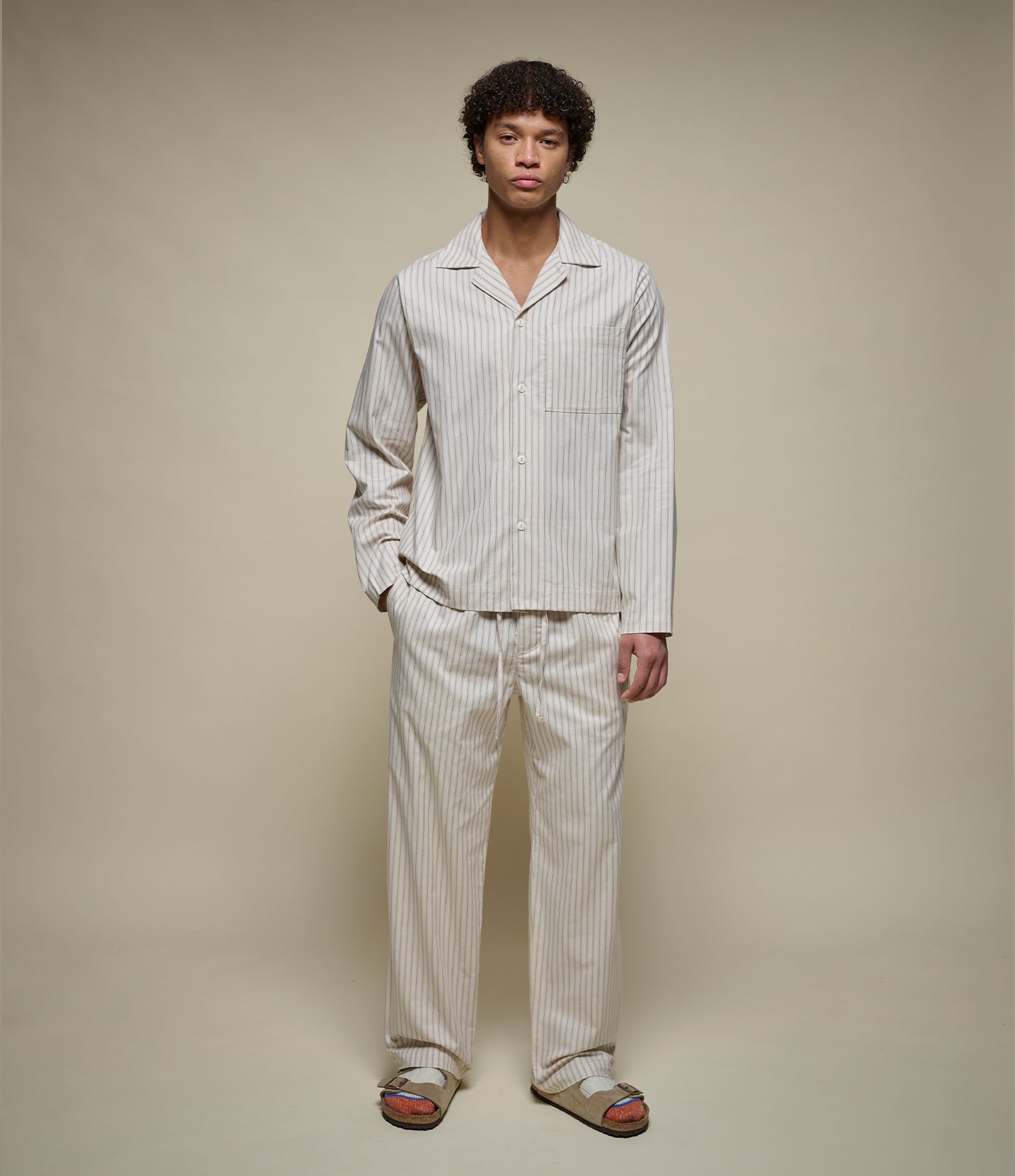 A man modeling a matching two-piece pajama set with vertical stripes, consisting of a long-sleeve button-up shirt and matching pants, paired with brown slide sandals against a solid beige studio background.