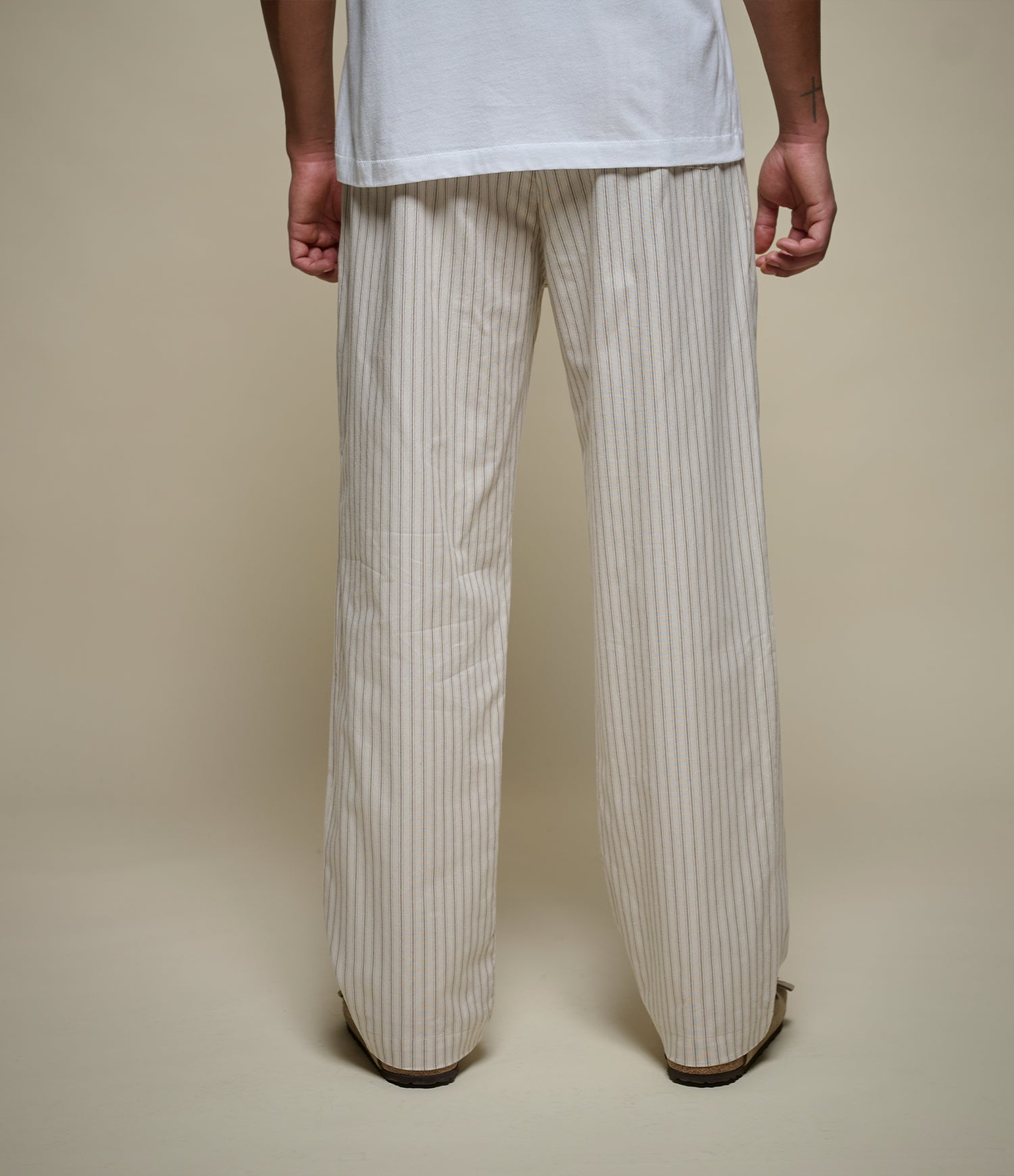 A person shown from the back wearing loose, wide-leg trousers with thin vertical beige and white stripes, paired with a white t-shirt, against a solid beige studio background.