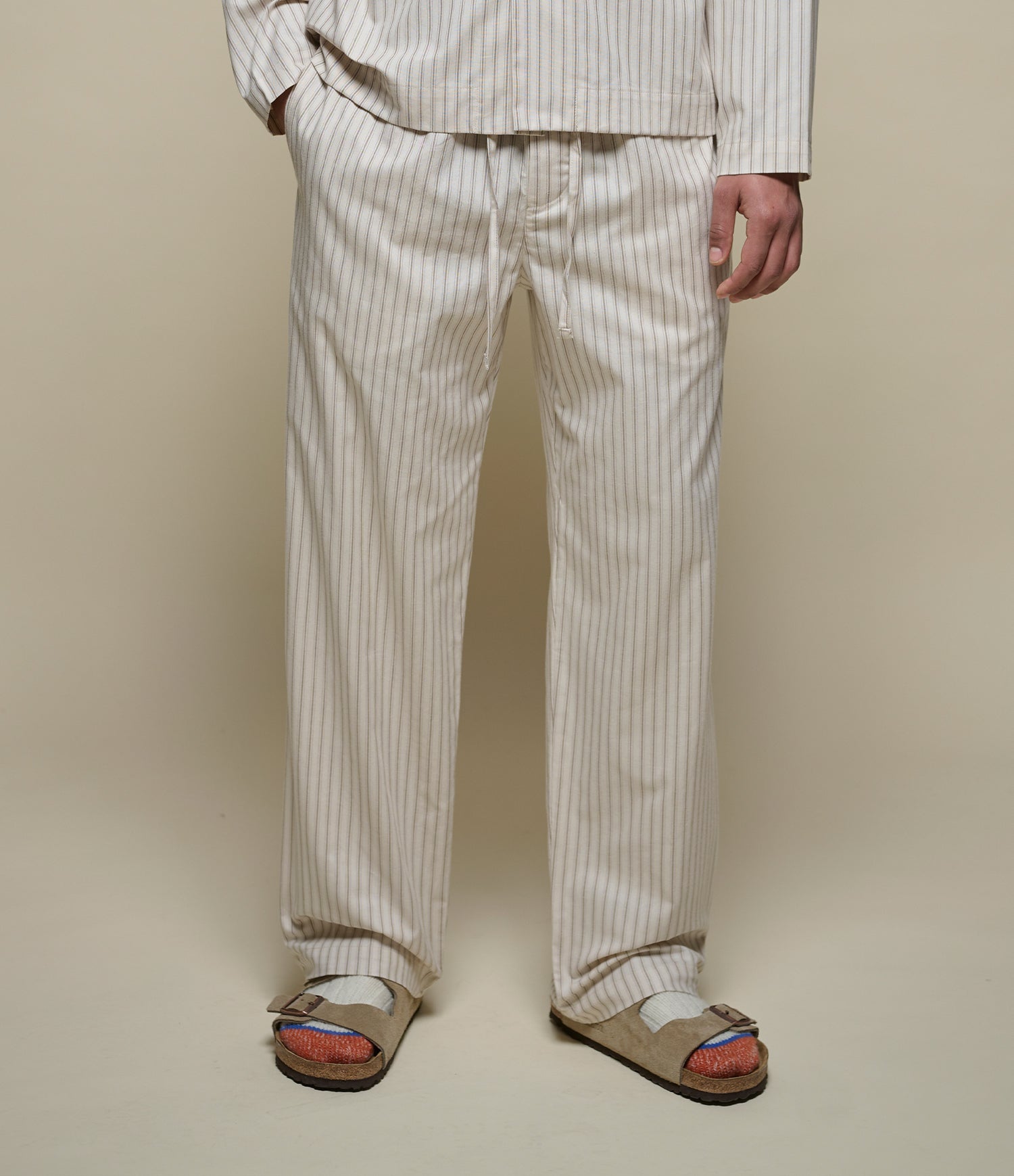 A person wearing cream and beige vertically striped pajama pants and a matching long-sleeve top, styled with tan slide sandals, against a solid beige studio background.