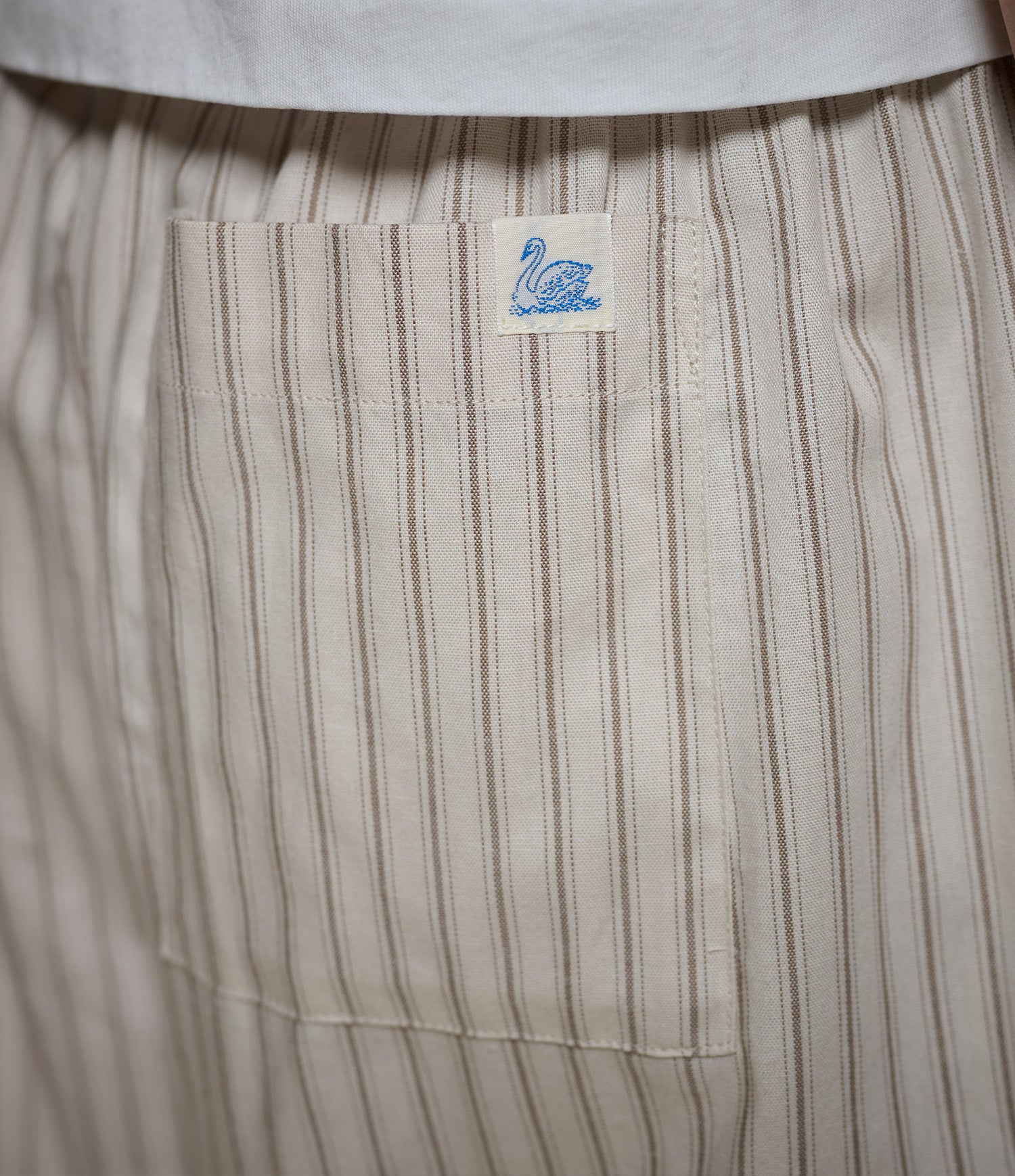 Close-up of a striped fabric garment, likely a shirt or pants, showing a beige pocket with a small blue swan logo patch.
