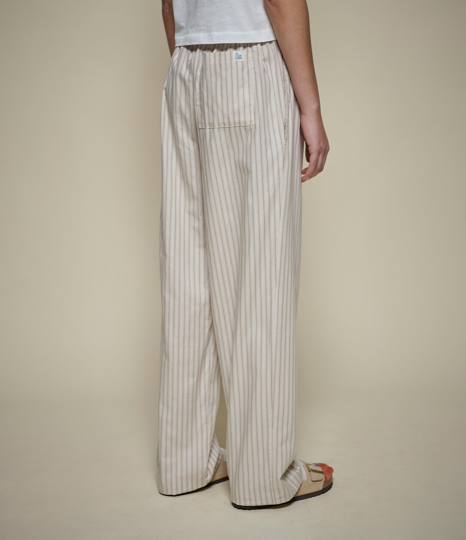 A person shown from the back wearing wide-leg striped pants, a white top, and flat sandals against a plain beige studio background.