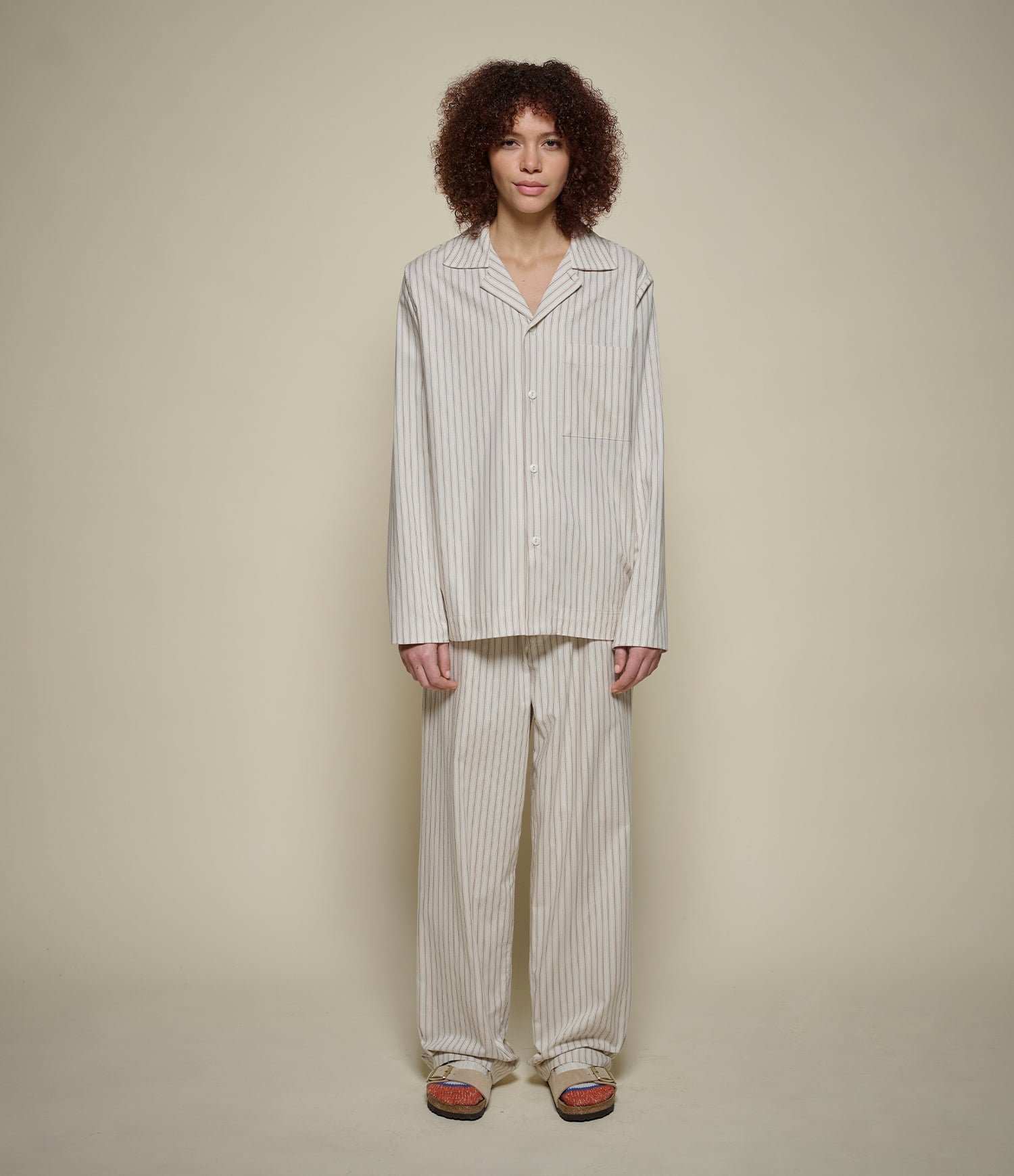 A model with curly dark hair stands against a plain beige studio background wearing a matching two-piece pajama set with vertical stripes. The set includes a long-sleeve button-front top and wide-leg pants in an off-white fabric with thin dark stripes, paired with brown slip-on shoes.