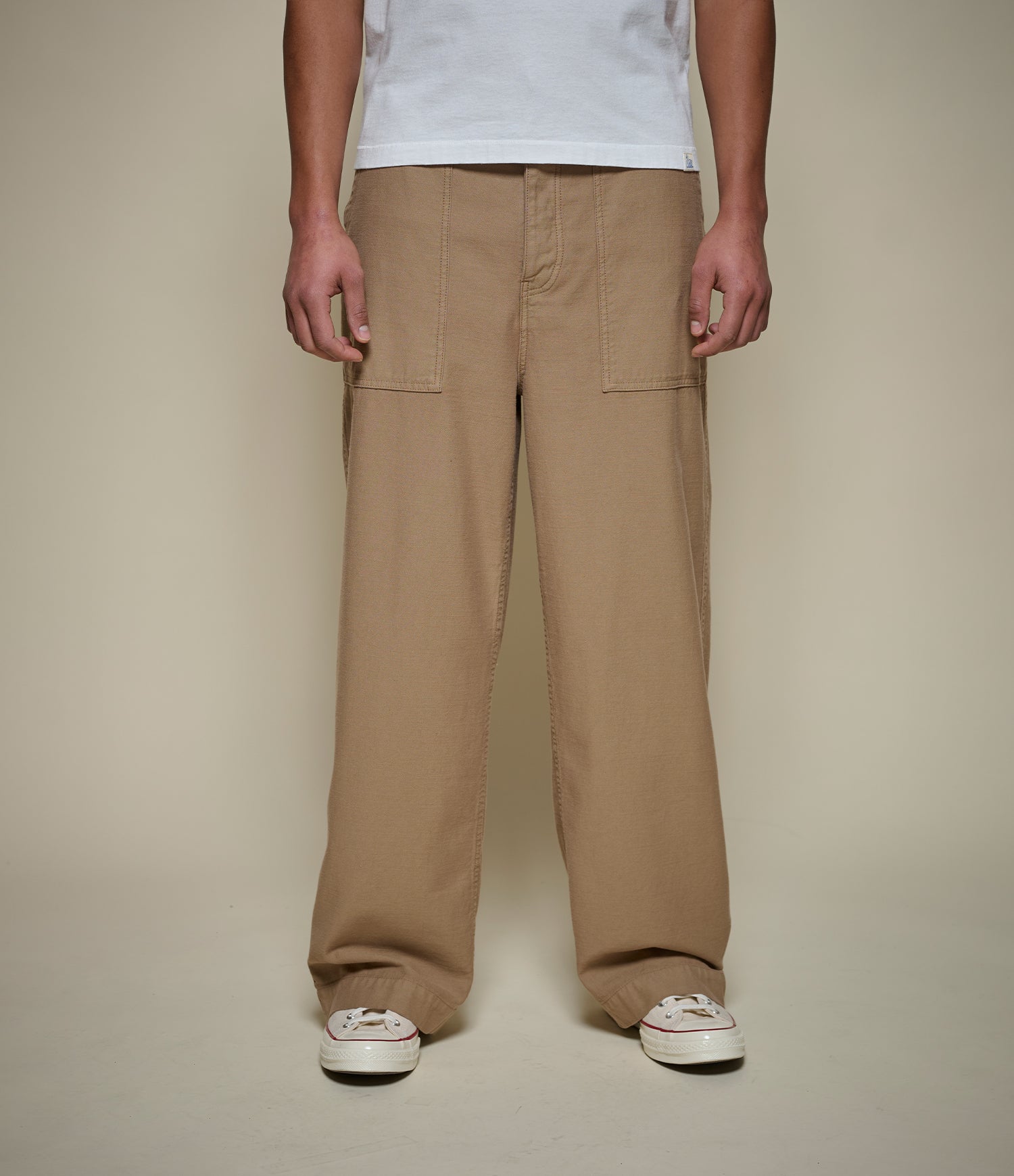 A person wearing loose-fit tan cargo pants with a white t-shirt and white sneakers, standing against a plain beige studio background.