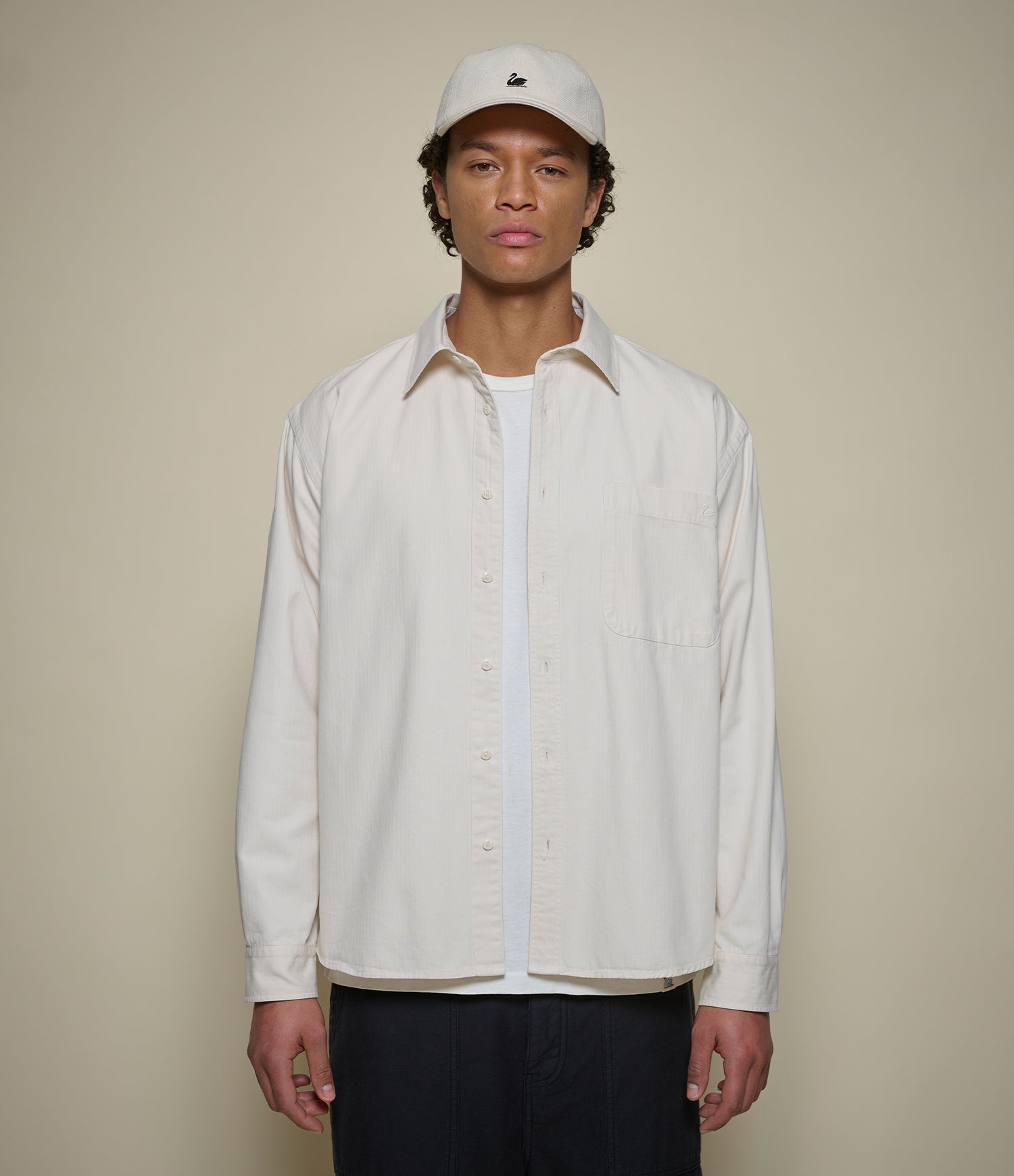 A man wearing a cream-colored long-sleeve button-up shirt over a white t-shirt, paired with a matching cream baseball cap and dark pants, standing against a solid beige background.