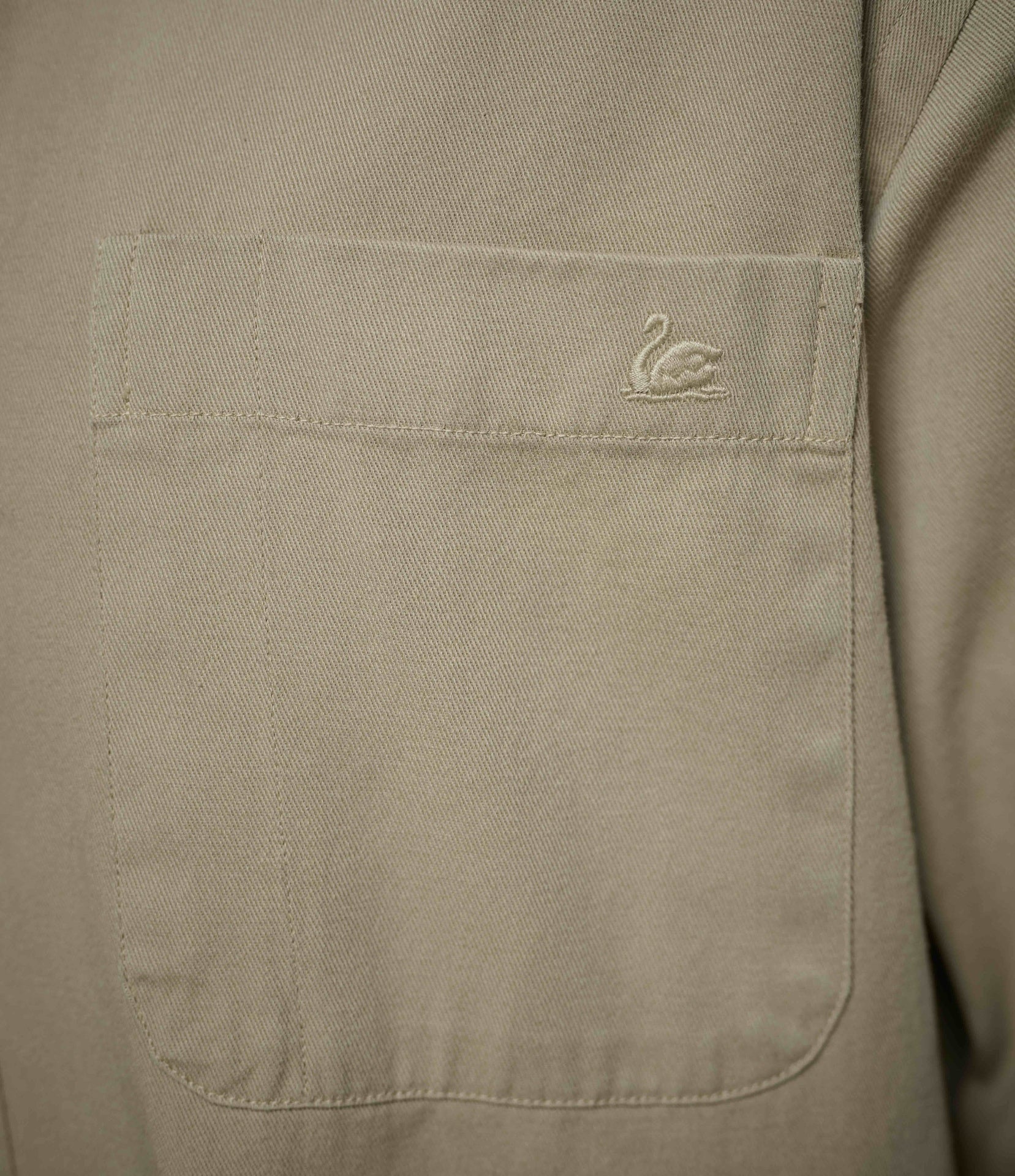 Close-up of a beige fabric garment showing a front patch pocket with a small embroidered swan logo on the upper right side of the pocket.