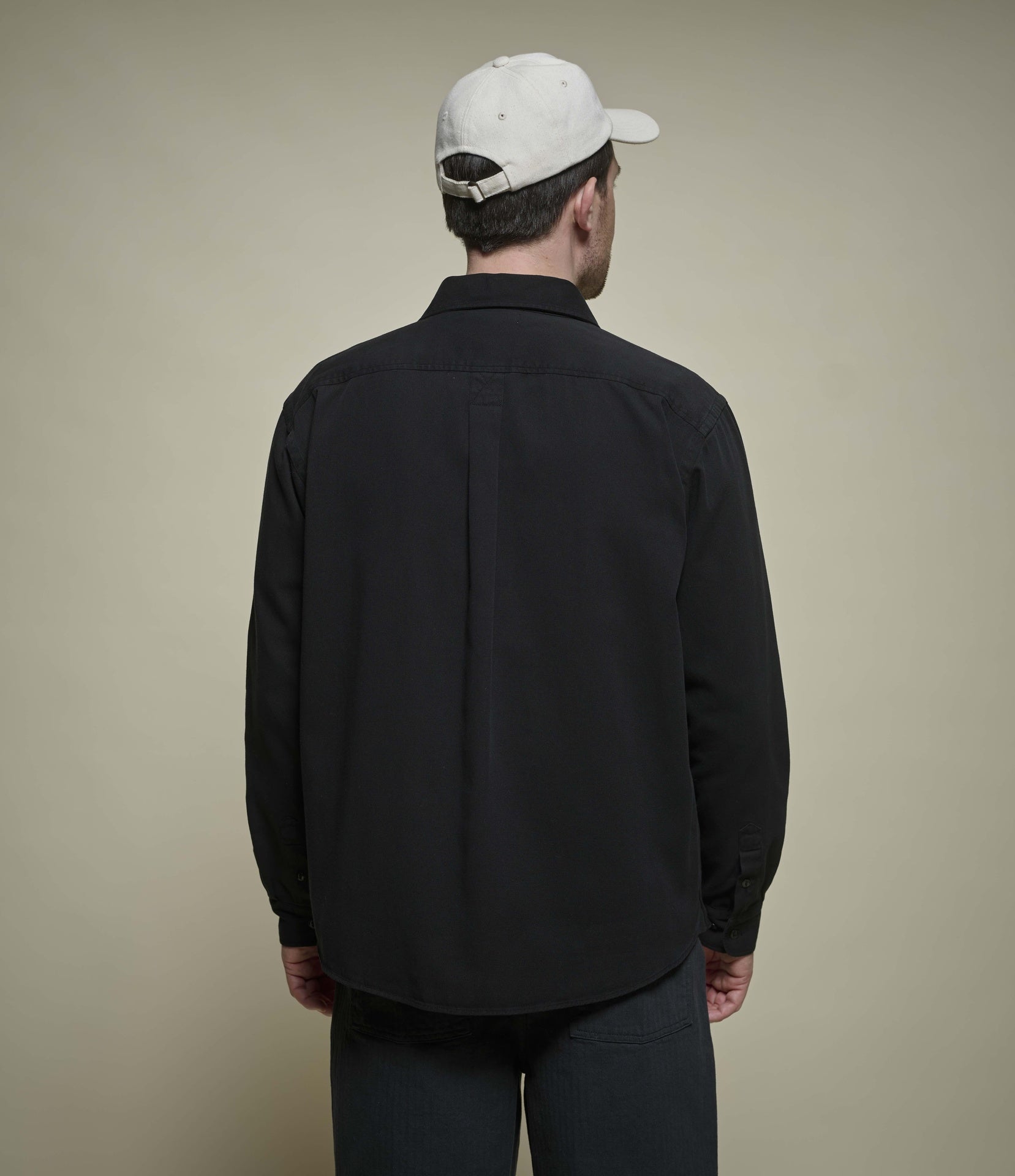 A man shown from the back wearing a black long-sleeve button-up shirt, dark trousers, and a white baseball cap against a plain beige studio background.