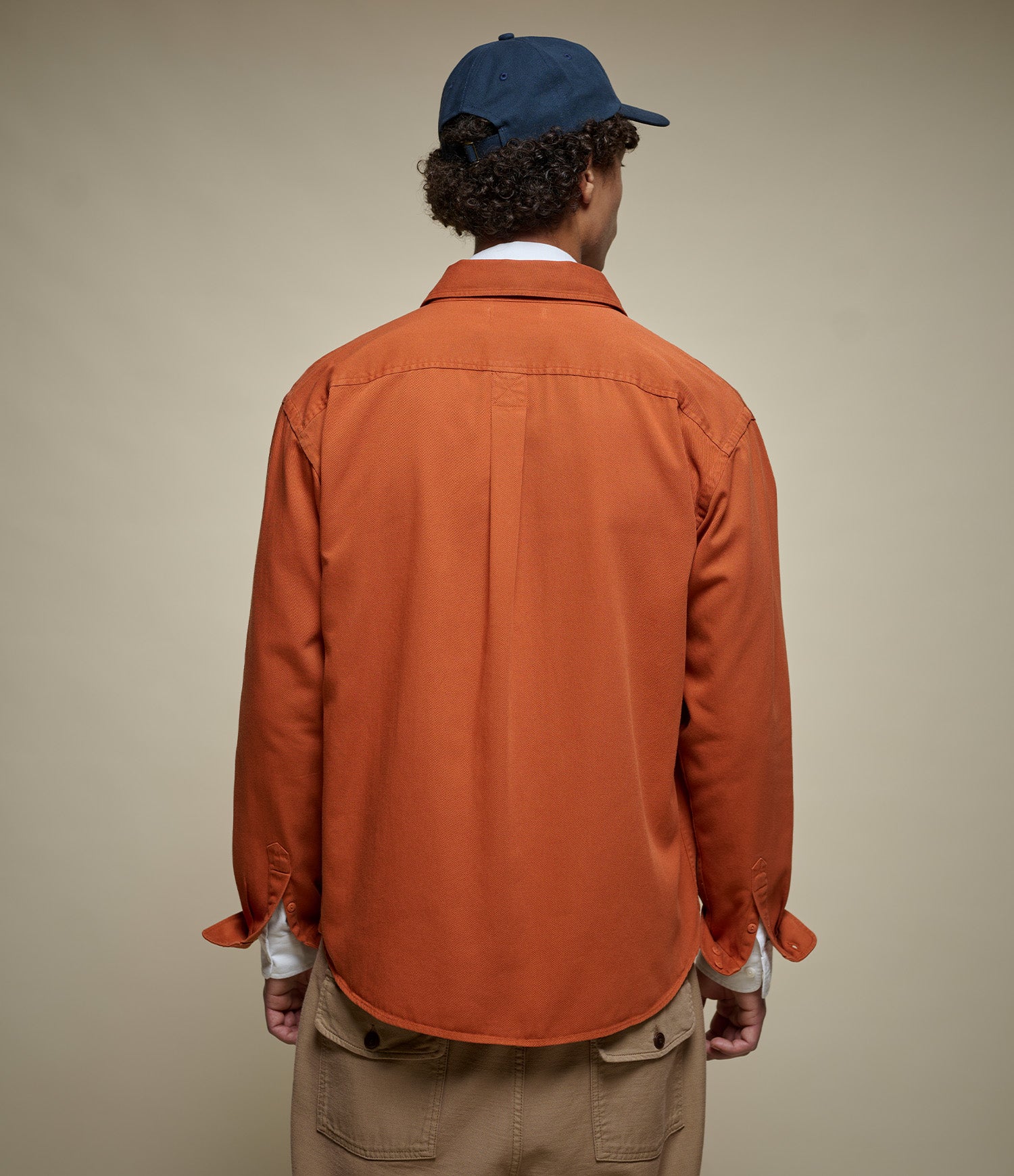 A man viewed from behind wearing a rust-orange long-sleeve button-up shirt, tan pants, and a navy blue baseball cap against a plain beige studio background.