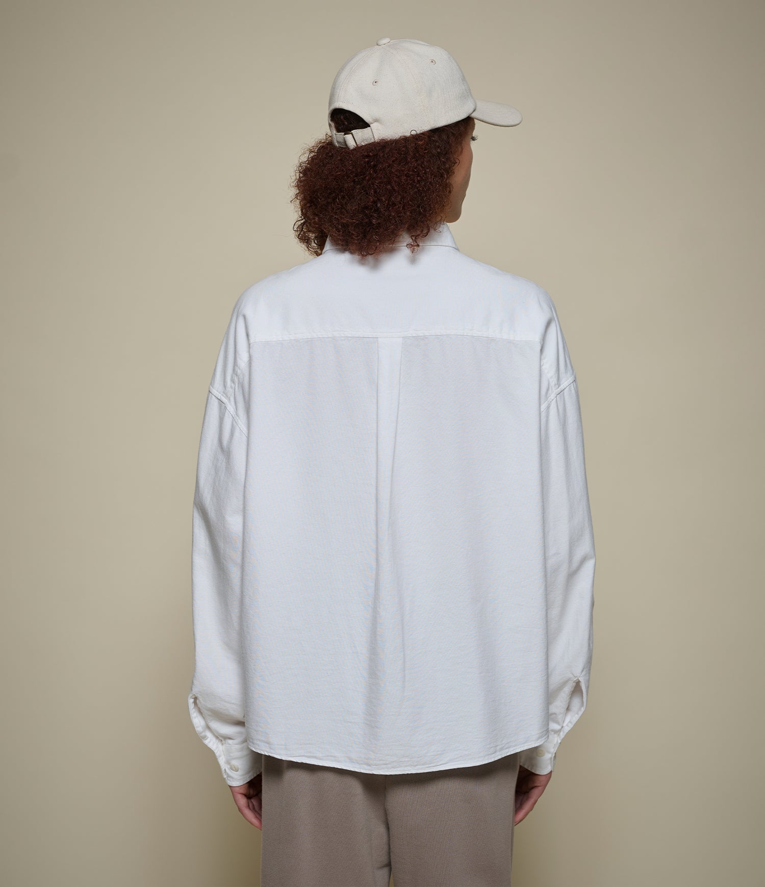 Person wearing a white cropped oxford shirt and beige cap from behind on a beige background
