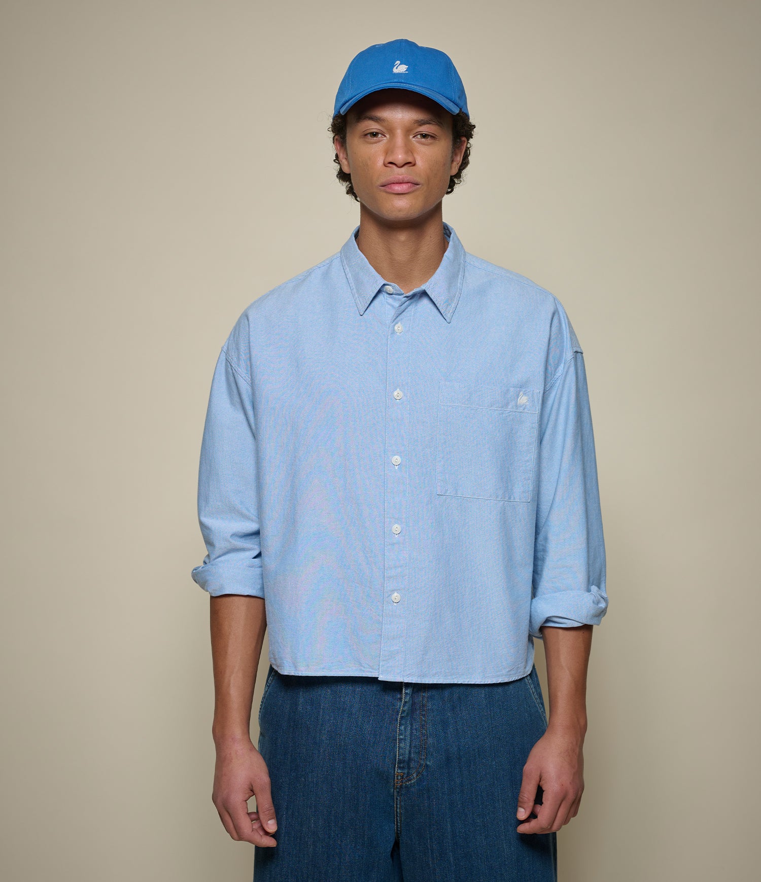 Man wearing a blue cropped oxford shirt and blue cap against a beige background